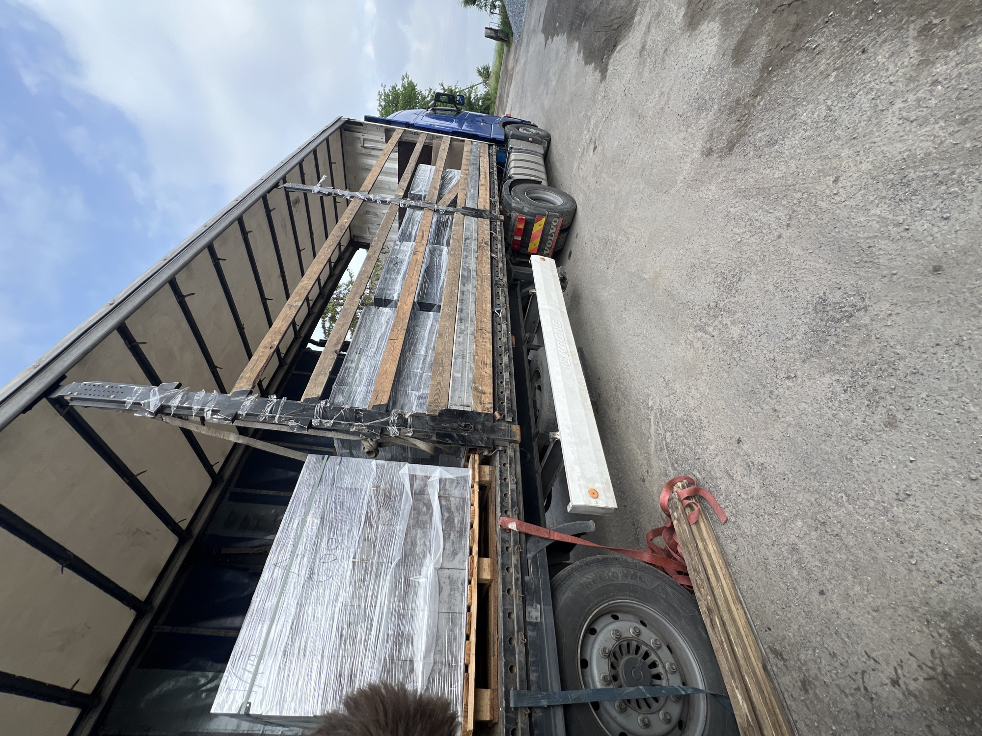A blue Volvo semi-truck with a curtain-sided trailer is parked on an asphalt surface under a partly cloudy sky. The image is captured from a low angle, looking up towards the side of the trailer.
Inside the open trailer, numerous rectangular packages, wrapped in clear or silvery plastic, are stacked on pallets. These packages appear to be a type of building material or insulation. Some illegible handwritten marks are visible on the wrapping of these items. Wooden beams are integrated into the internal structure of the trailer, possibly serving as load securing elements or part of the cargo itself.
Attached to the trailer's chassis is a white, elongated storage box with the brand name "KRONE" clearly visible on its side. A spare tire, marked "PEGASUS" on its sidewall, is mounted externally to the trailer, secured by straps. Red tie-down straps are seen near the spare tire and draped over a bundle of wooden planks lying on the ground next to the truck.
The blue cab of the Volvo truck is partially visible towards the front, with the "VOLVO" logo displayed prominently on its rear. In the background, green trees and distant foliage are visible. The ground is a rough, grey asphalt or concrete. The scene takes place during daytime in an outdoor industrial or storage area, in Uhry, Ukraine.