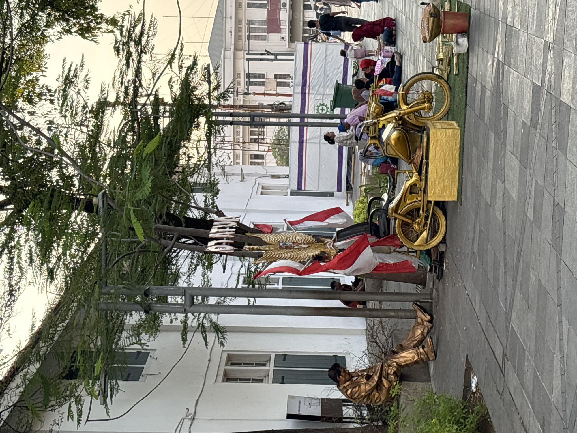 In the Kota Tua area of West Jakarta, Indonesia, a street performer dressed in gold is seated on a ledge near a white building with green-shuttered windows. In front of the building, a golden motorcycle is displayed on a patch of artificial grass, with its base covered in gold-painted bricks. To the right of the motorcycle are various golden objects, including what appears to be a small grill.
Several people are gathered in the background, some sitting on the ground and others standing. Among them, a woman in a plaid shirt and a blue headscarf is seated, while a child in a pink outfit walks past. Another group of people, including women in headscarves, are seated on the ground, some appearing to be tourists. A man in a black shirt is walking away from the scene.
Red and white Indonesian flags are displayed prominently on poles, along with a golden eagle emblem. A sign is visible on a tree in the background, though the text is partially obscured. The overall scene depicts a public space with street art, performers, and onlookers, characteristic of a tourist attraction.