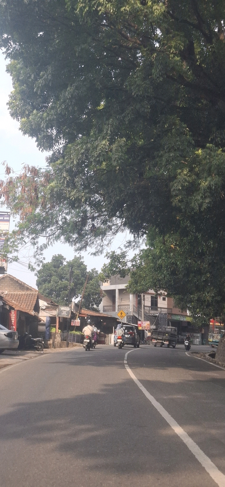 The image depicts a street scene in Cilaku, Indonesia, under a bright sky, possibly in the morning or afternoon given the shadows cast by trees. The road is paved and curves to the right. On the left side of the road, there are several small buildings with traditional tiled roofs, and some modern structures are visible further down the street. A large tree with dense foliage dominates the upper right portion of the image.
Traffic is present on the road, including several motorcycles and at least one car. On the leftmost side, a silver car is parked. A few motorcycles are in motion, with riders visible. One motorcycle is being ridden by a person wearing a light-colored shirt and dark pants. Another motorcycle has two people on it, one wearing a hijab. A black car is also moving on the road, and a yellow pedestrian crossing sign is visible on the right side of the road. Further down the road, a truck is also present.
Various signs are visible on the buildings, including one with the text "ENGKELMO" and another with "10000". A sign with "MIE BANSO" is also visible on a building on the right. The overall impression is a bustling street with a mix of residential and commercial establishments, common in Indonesian towns.