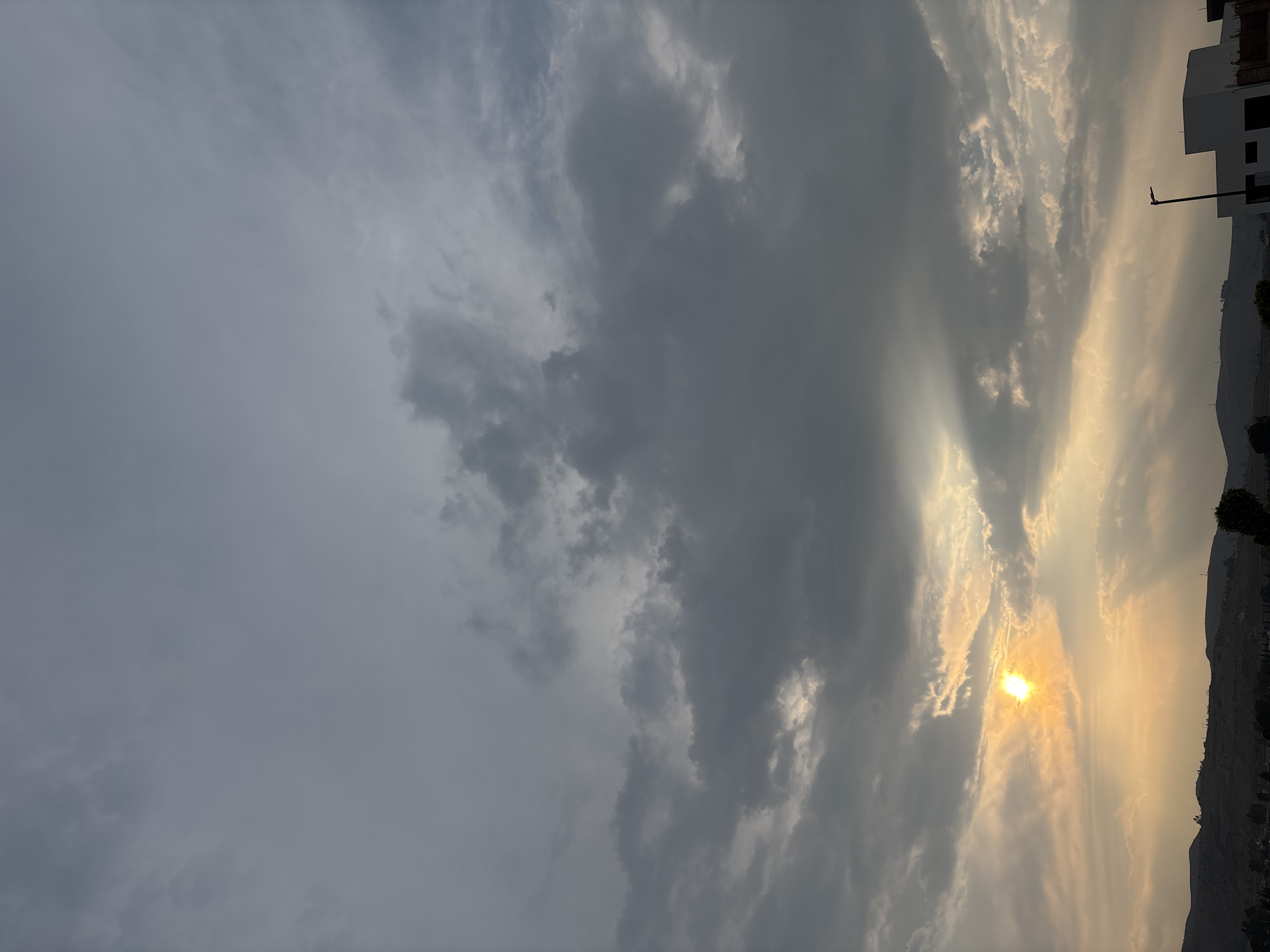 The image depicts a sky filled with dark gray clouds and a bright sun, possibly at sunrise or sunset. The sun's rays are visible through openings in the clouds. The background shows a portion of a building or structure and what appears to be a mountainous or hilly landscape. It's difficult to confirm the location in Santa Clara Ocoyucan, Mexico, without more local landmarks visible in the frame.