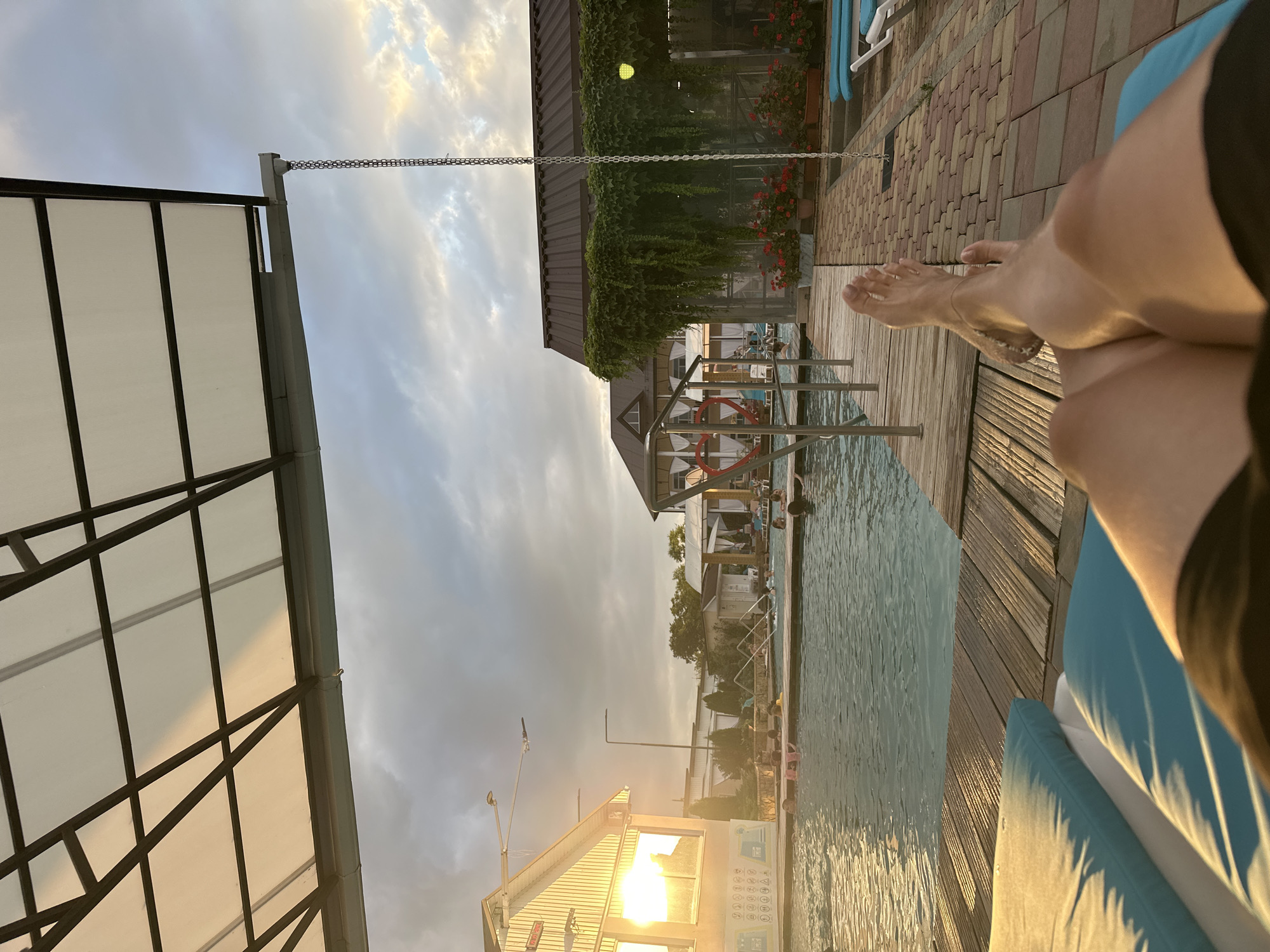 The image depicts a poolside scene with a person's legs and feet visible in the foreground, resting on a blue chaise lounge. The setting appears to be a resort or spa, likely in Yaroslavskaya, Russia, given the context. The sky is cloudy with hints of sunset colors. The pool itself is visible, with several people swimming and relaxing. A wooden deck surrounds the pool, and beyond that, there are buildings, one of which has ivy growing over its roof. The overall atmosphere is one of leisure and relaxation.