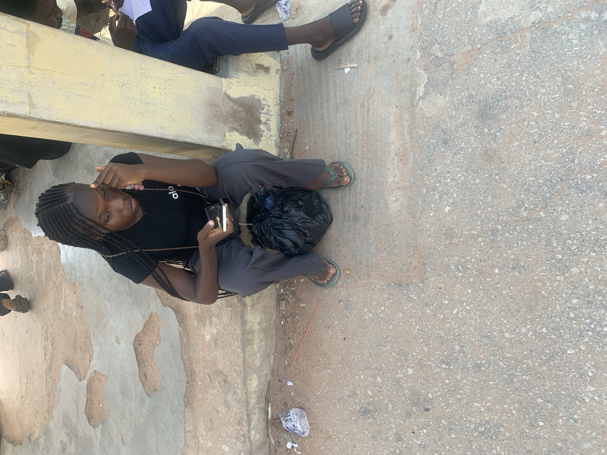 A young Black female with braided hair is sitting on a concrete ledge in Jalingo, Nigeria. She is wearing a black t-shirt and gray pants, with green flip-flops on her feet. She is holding a smartphone and appears to be looking at it, with her finger touching her forehead. Next to her is a black plastic bag. To her right, another person is sitting, wearing dark blue pants and black flip-flops, and appears to be writing on a piece of paper. The environment is an outdoor area with cracked concrete and sandy ground. There is some litter on the ground, including a clear plastic bag and a red stick. A light yellow concrete pillar is also visible to the right of the young woman.