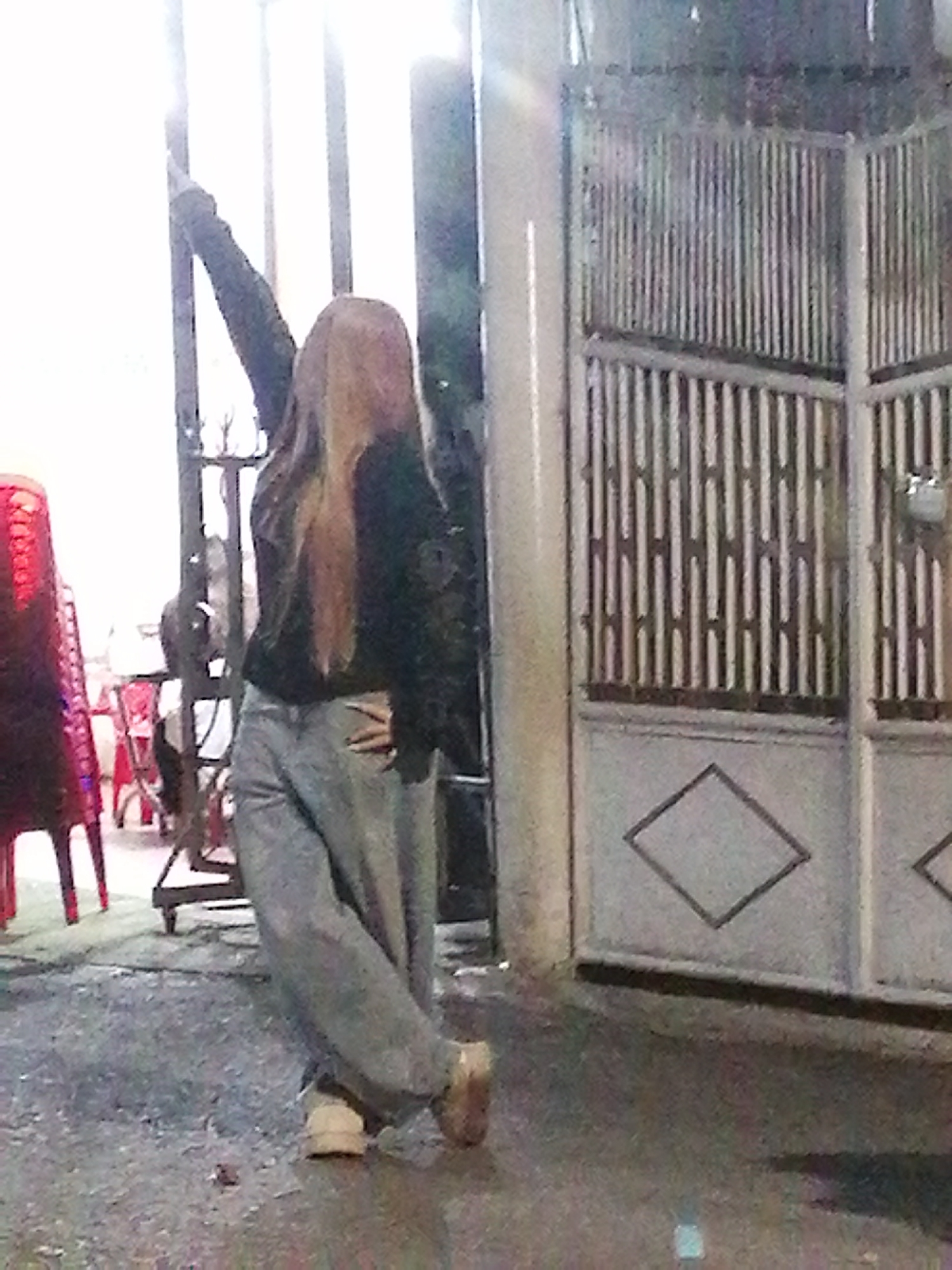 A young woman with long, blonde hair and a blurred face stands on a wet, paved outdoor area, leaning against a pillar and gesturing with her left arm. She is wearing a dark jacket and loose-fitting light-colored pants. In the background, a large, ornate metal gate with vertical bars and a diamond pattern is visible, partially obscuring a building. To the left of the woman, red plastic chairs are stacked. The scene appears to be a street or alleyway in Da Lat, Vietnam, likely at night or in low light conditions, given the reflections on the wet ground and the bright light source behind the woman.