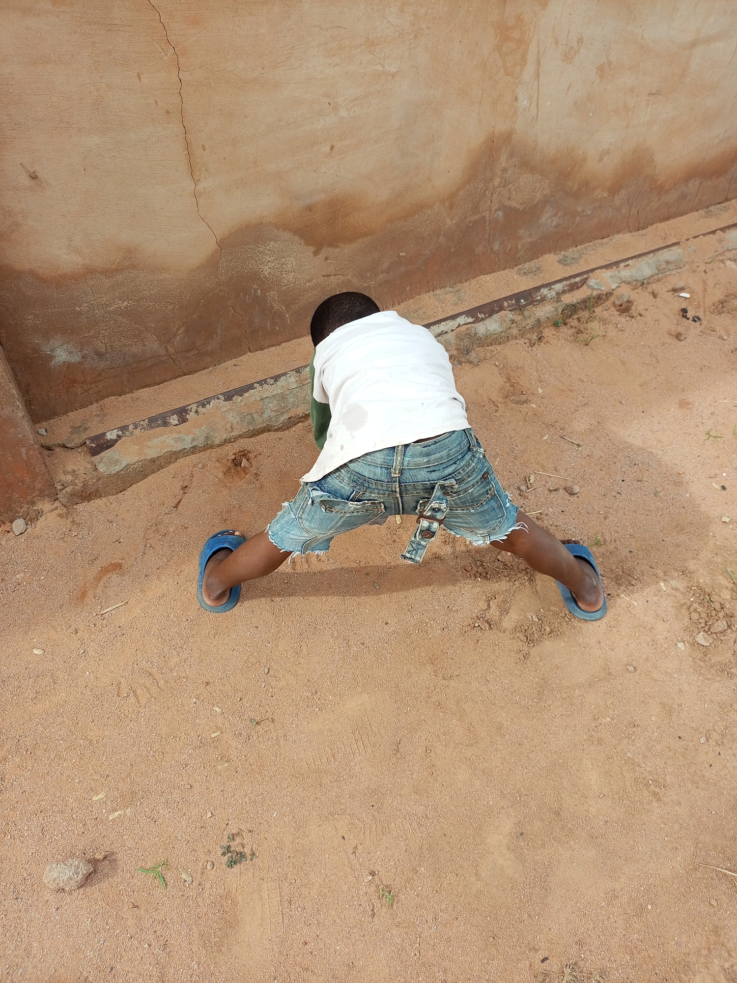 A young boy is on his hands and knees on sandy ground, facing away from the camera. He is wearing a white t-shirt and distressed denim shorts with blue flip-flops. He is positioned in front of a weathered, light brown wall with visible cracks and discoloration. A metal bar runs along the base of the wall. The ground is covered in sand with some small stones and sparse vegetation. The scene appears to be outdoors, possibly in a courtyard or along a building exterior.