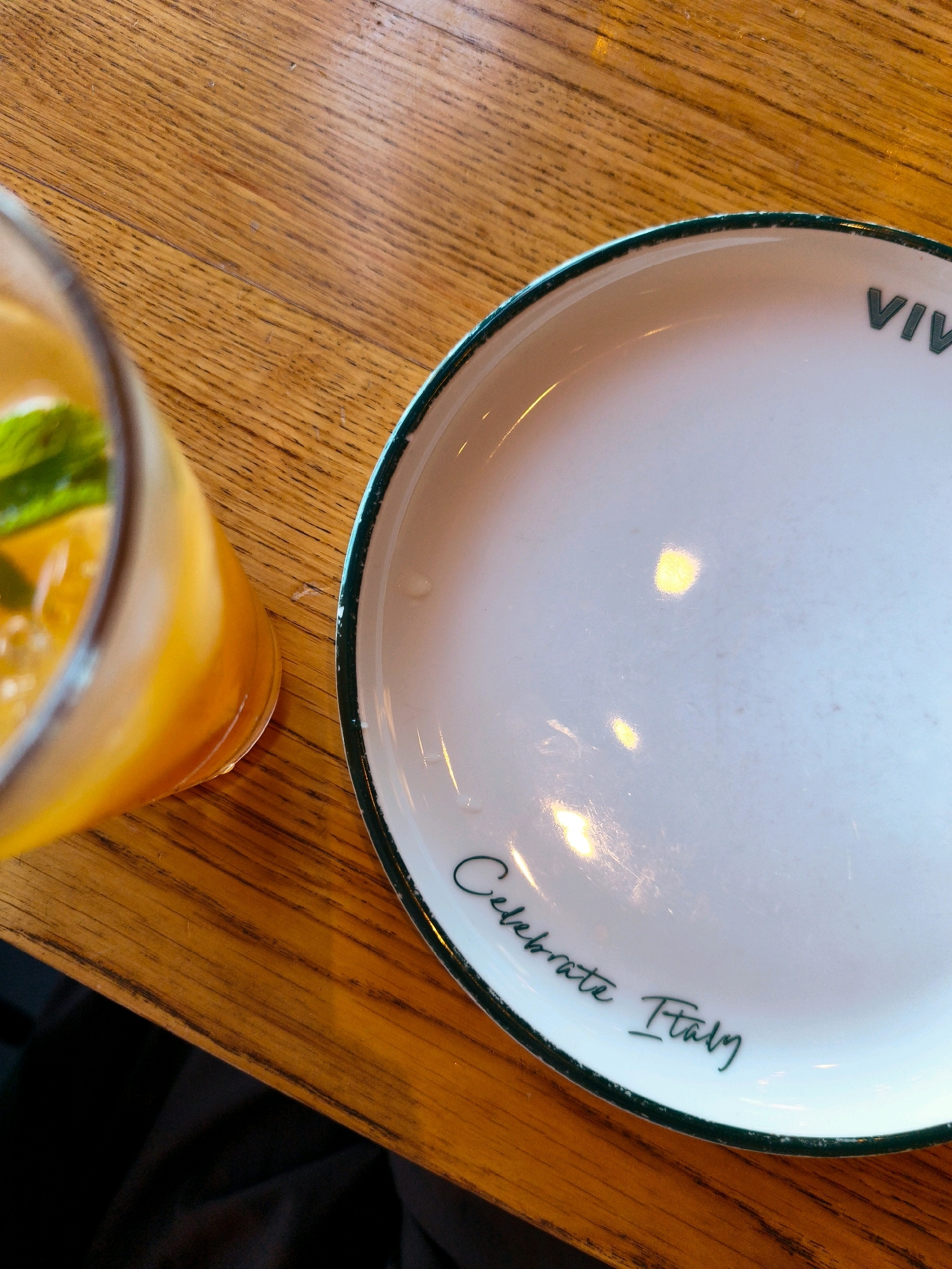 The image shows a close-up view of a wooden table with a clear glass of orange beverage and a white plate with a green rim. The beverage appears to be a juice or cocktail, with some mint leaves floating in it. The plate has a green border and text that reads "Celebrate Italy" in a stylized font. Above that, also in green, is the partial word "VIV". The lighting suggests it is daytime, possibly in a restaurant or cafe. No people are visible in the image. The location is described as Netivot, Israel.