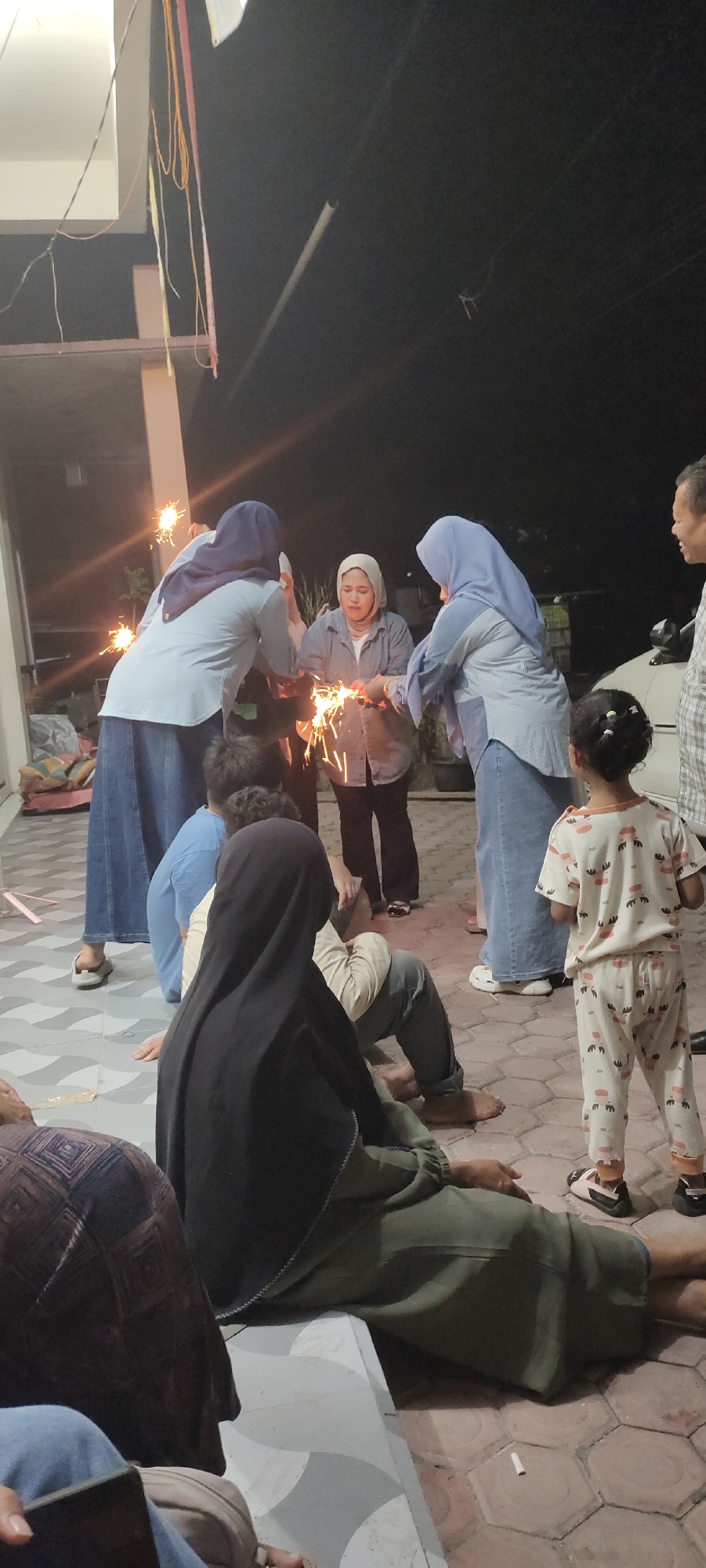 The image shows a group of people in Bireuenmeunasahdayah, Indonesia, at night. Three women are standing and holding sparklers. One woman is wearing a light gray shirt and white hijab. The other two women are wearing headscarves. A little girl is standing to the right, wearing pajamas with a pattern and shoes. Several people are seated on the ground; one woman in the foreground is wearing a black hijab. The ground is paved with hexagonal tiles. In the background, there is a building with a porch. A car is parked on the right. Several pieces of string are hanging near the building.