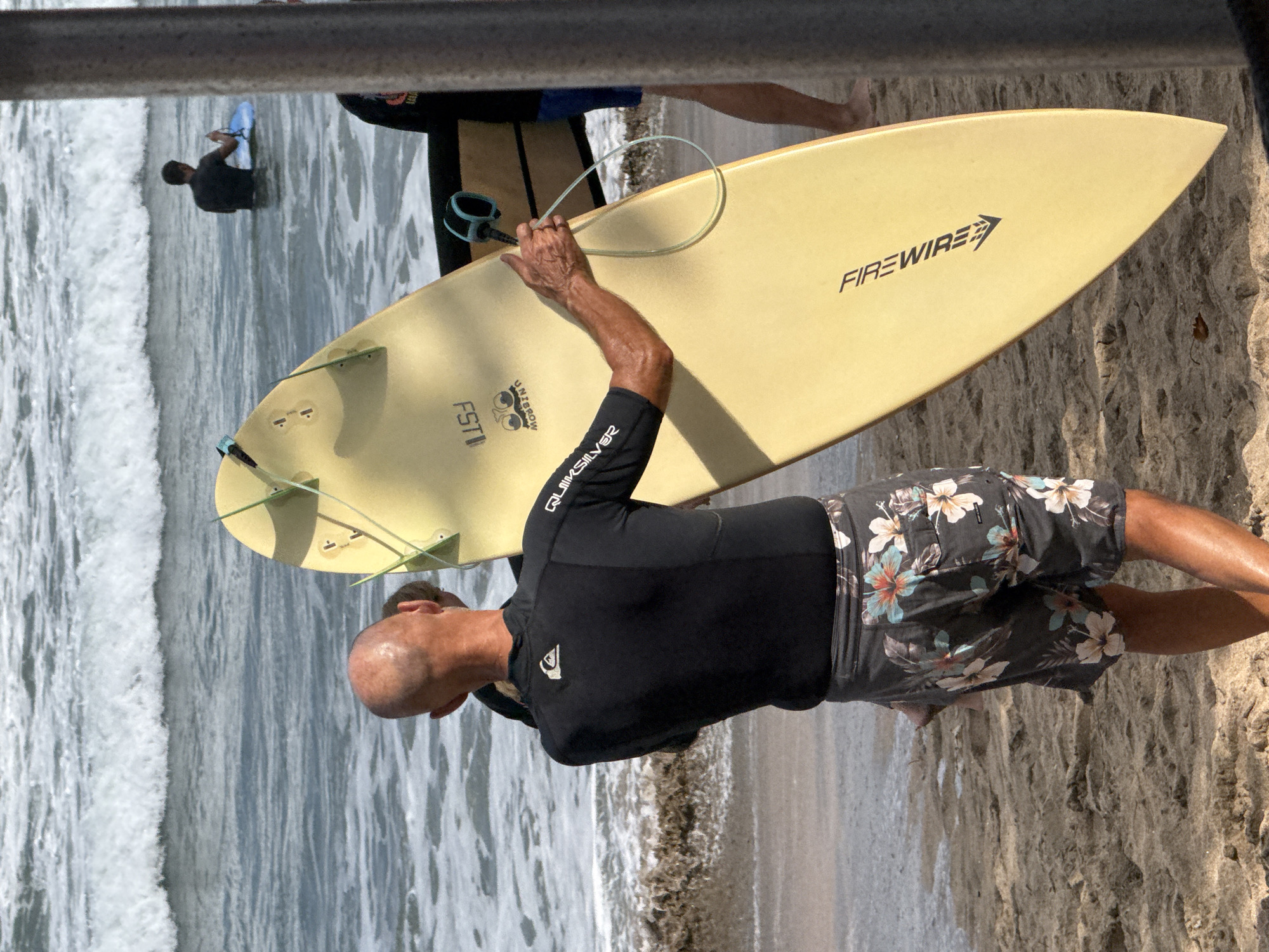 A man with a shaved head and tanned skin is standing on a sandy beach in Kuta, Indonesia. He is wearing a black wetsuit and floral-patterned board shorts. He is holding a pale yellow surfboard, the leash of which is attached to his wrist. The surfboard is positioned vertically in front of him. In the background, the ocean has waves, and another person is visible in the water holding a blue object. The environment is a beach with sand in the foreground and the ocean in the background.