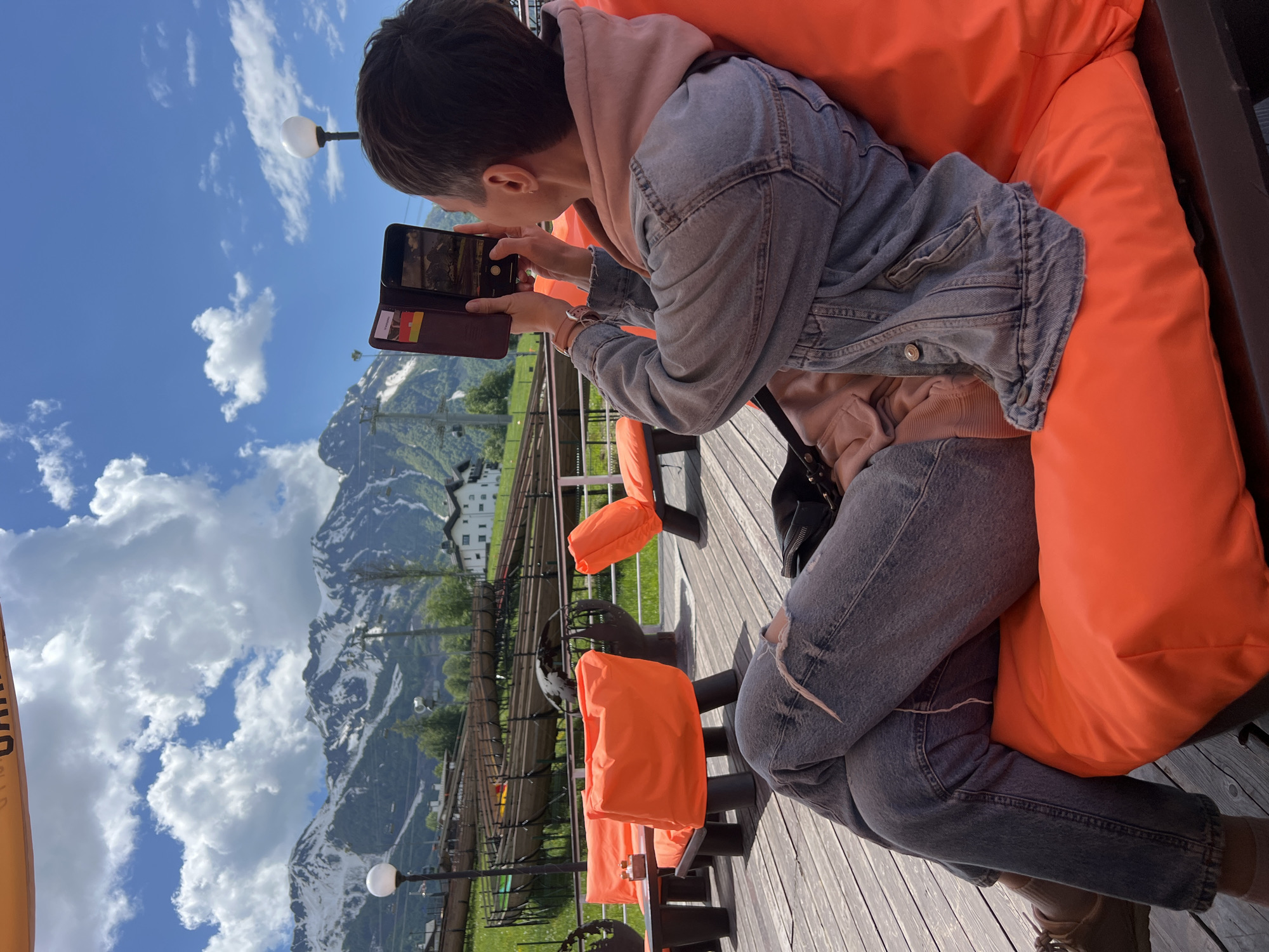 The image captures a person seated on a bright orange chair or cushion on an outdoor wooden deck, actively photographing a scenic mountain panorama with their smartphone. The individual, seen from a side and slightly rear angle, has short dark hair and is dressed in a light pink or beige hooded sweatshirt layered under a denim jacket, grey ripped jeans, and light brown shoes. A watch is visible on their left wrist.
The person is holding a black smartphone, housed in a brown wallet-style case, with both hands to frame the shot. The phone screen displays a digital rendition of the mountain landscape, confirming they are capturing the view. Visible within the wallet case are a few small, colorful rectangular items, likely cards.
The background presents a breathtaking vista of towering, snow-capped mountains under a brilliant blue sky, adorned with scattered white clouds. The lower slopes are green, and several buildings, characteristic of a mountain resort, are visible among the trees. A structure resembling a mountain coaster track or similar leisure facility can be seen traversing the landscape. The wooden deck features other unoccupied orange chairs and tables, along with lampposts, creating a relaxed outdoor seating area.
The scene takes place during the daytime, under clear and sunny weather conditions, suggesting a pleasant environment. The activity implies a leisurely visit or vacation in a picturesque mountain region, consistent with the provided location context of Estosadok, Russia. To the far left, a partial, indistinct word or logo is visible on an orange/yellow banner.