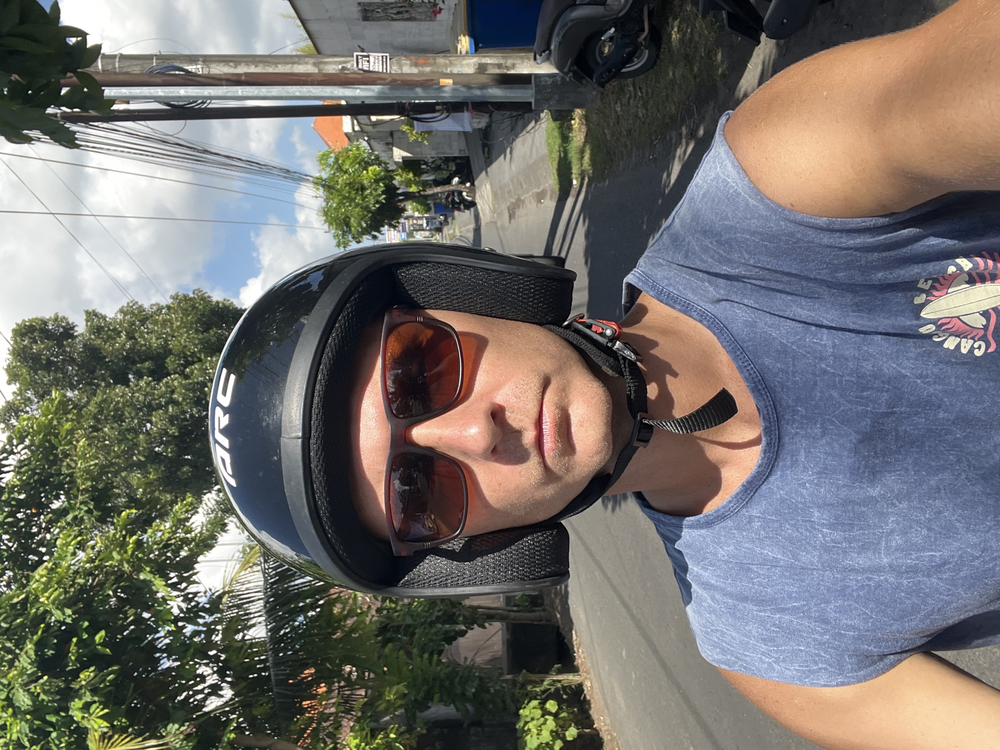 A man wearing a black motorcycle helmet with "ARC" written on it and brown sunglasses is looking forward. He is wearing a blue sleeveless t-shirt with a graphic of a surfboard. The man appears to be riding a motorcycle down a paved road in a tropical environment. Trees line the left side of the road, and buildings and more trees are visible on the right. There are power lines overhead and a parked motorcycle on the right side of the road. The sky is blue with white clouds. The scene appears to be located in North Kuta, Indonesia, given the architecture, vegetation, and presence of motorcycles.