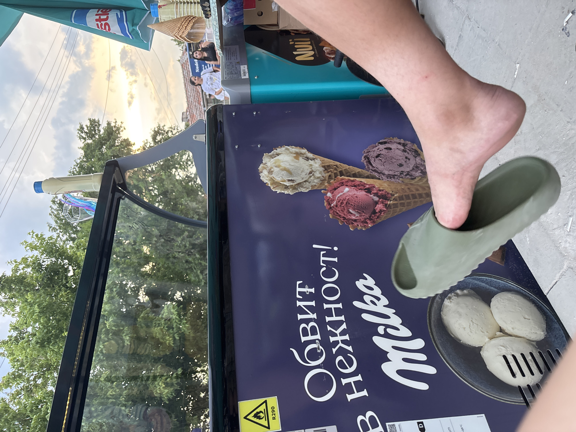 The image depicts an outdoor ice cream vendor's setup in Kostievo, Bulgaria, captured from a low perspective. In the immediate foreground, a light-skinned bare left foot, partially inserted into a dull green slide sandal, rests on a concrete surface. The central object is a purple-paneled ice cream freezer displaying prominent white "Milka" branding and the Bulgarian text "ОБВИТ В НЕЖНОСТ!" (WRAPPED IN TENDERNESS!). The panel features images of three ice cream cones with different flavored scoops (vanilla, berry, and chocolate) and three scoops of white ice cream in a blue bowl with a serving utensil. A yellow triangular warning label with a flame symbol and "R290" is affixed to the upper left of the freezer.
Above the freezer, a glass barrier reflects the dense green canopy of trees against a light blue sky with a distinct orange and yellow sunset glow visible on the right horizon. To the right of the main freezer, a teal-colored upright freezer with "Nuii" branding is partially visible, alongside an aqua-colored Nestlé branded umbrella. An array of empty waffle cones, paper cups, and assorted plastic utensils are arranged on a counter surface. On top of the freezer structure, a beige squeeze bottle with a blue cap and a clear container holding colorful plastic cutlery are present.
In the mid-ground, behind the vendor's setup, two women are observable; one in a white graphic t-shirt looking left, and another in a dark top looking right. They stand near a blue vehicle and a building with a red tiled roof, silhouetted against the sunset. The ground beneath the foot and vendor's station is light gray concrete, showing some scuffs and marks.