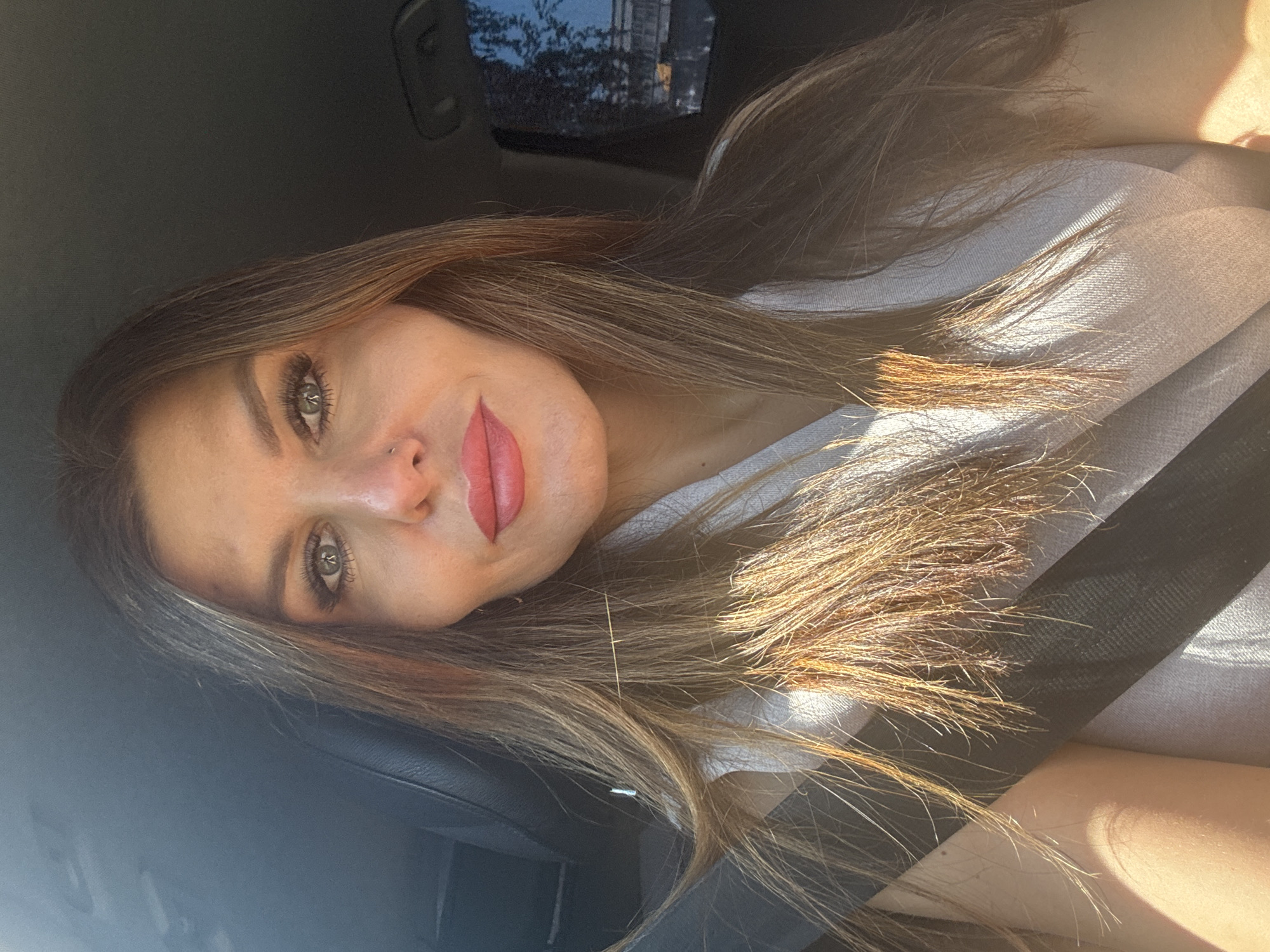 A woman is seated in a car, wearing a light-colored shirt with a dark seatbelt. The image is rotated, making it appear as though she's lying down. She has long, light brown hair with highlights and is wearing red lipstick. Sunlight illuminates part of her face and hair. Outside the car window, there is a blurred view of buildings and trees, indicating an urban environment. There is no specific indication that this is in Chaikovsky, Russia.
