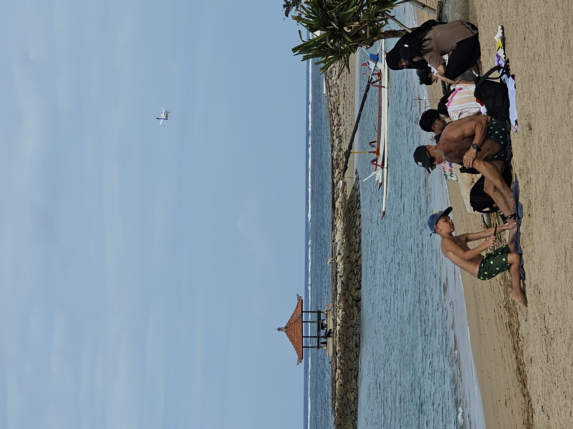 The image depicts a beach scene in Denpasar, Indonesia, under a partly cloudy sky. In the foreground, a group of people are relaxing on the sand. A man in a black baseball cap and sunglasses is sitting with his legs extended, wearing patterned swim shorts. Beside him, a young boy wearing a blue baseball cap and green patterned shorts is kneeling on the sand, appearing to be engaged with something on the ground. Behind them, several other individuals are seated or leaning, including women wearing headscarves and casual attire, and a child. Several bags are scattered on the beach around the group.
In the middle ground, the turquoise ocean meets the sandy shore. A traditional Indonesian outrigger boat, painted white with colorful accents, is moored in the shallow water. Further out, a man-made structure with a red-tiled, octagonal roof sits atop a stone jetty that extends into the sea. The water exhibits gentle waves.
In the background, the horizon is marked by a thin white line of breaking waves, suggesting a reef or shallow area offshore. High in the pale blue sky, a small, white airplane with visible landing gear is captured in flight. The overall scene conveys a relaxed atmosphere of a beach day in Denpasar.