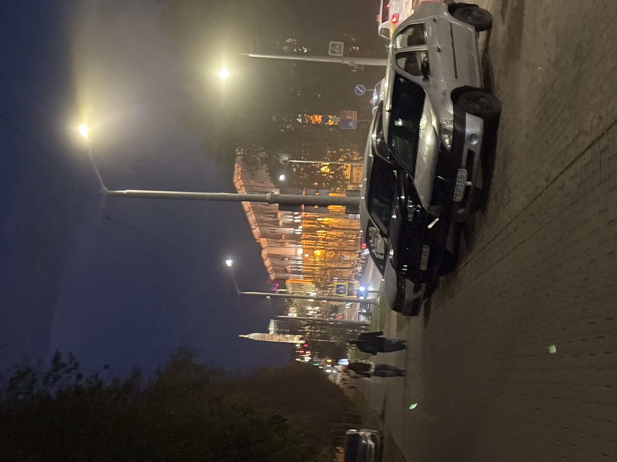 The image depicts a nighttime scene in Minsk, Belarus, along an urban street. A well-lit building with ornate architecture and glowing windows lines the right side of the street. The street itself is lined with parked cars, primarily sedans, with their headlights and taillights illuminated. A lone street lamp casts a warm glow over the pavement and the surrounding area, creating a somewhat hazy atmosphere. On the left side of the street, a sidewalk is visible, with two individuals walking away from the camera. Trees with dark foliage border the sidewalk. In the background, a tall, slender tower-like structure with illuminated windows stands out against the dark sky. The overall impression is that of a quiet, European city street at night, with a mix of modern and historical elements.