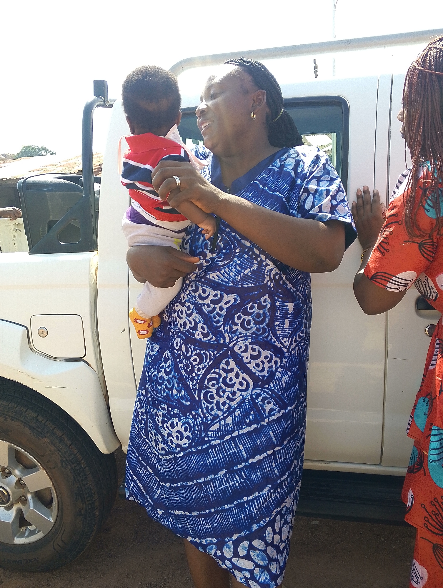 In an outdoor setting in Jos, Nigeria, a woman in a blue and white patterned dress stands next to a white pickup truck, holding a baby wearing a striped shirt and orange socks. The woman is smiling and looking at the baby. Another woman, wearing a red patterned dress, stands next to the truck, facing the first woman. The truck's tire and wheel are visible in the foreground. The background includes hints of buildings and foliage under a bright sky.