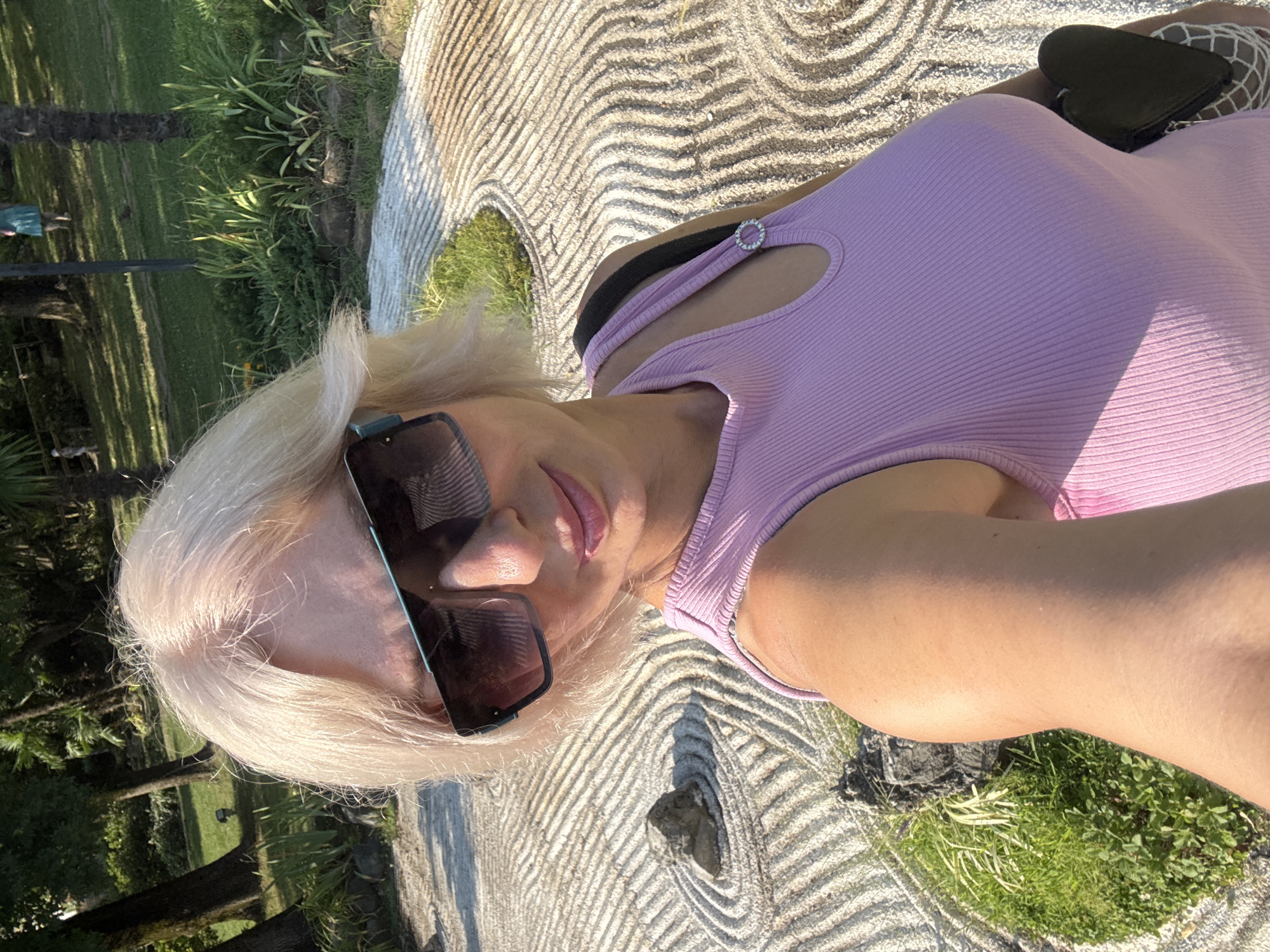 A blonde woman with short, light-colored hair and large sunglasses poses for a selfie in a Japanese garden. She is wearing a light pink ribbed tank top and carrying a dark bag. The background features a raked gravel garden with concentric circles and a large rock. Trees and lush greenery are visible in the distance, suggesting a park or botanical garden setting. The woman is looking at the camera with a slight smile. The scene appears to be outdoors in daylight. The location is identified as a Japanese garden in Sochi, Russia.