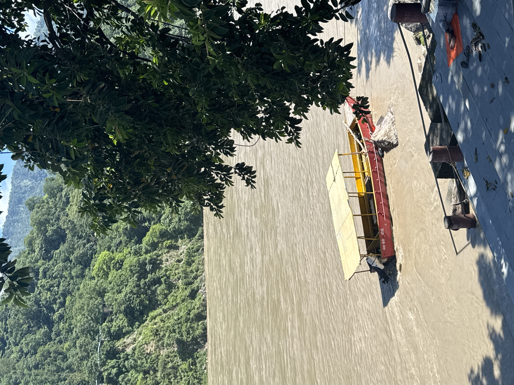 The image depicts a wide, muddy river flowing through a valley with steep, tree-covered hillsides in the background. In the foreground, a red and yellow boat with a canopy is partially submerged in the river, appearing to be either beached or caught on something underwater. A person is lying on an orange mat on stone steps near the riverbank, partially obscured by tree shadows. There are several bollards and a rope on the steps. The overall scene suggests a natural, somewhat rustic environment, likely a river ghat in Rishikesh, India, known for its spiritual sites and proximity to the Himalayas. The lighting indicates it is daytime, with the sun casting strong shadows.