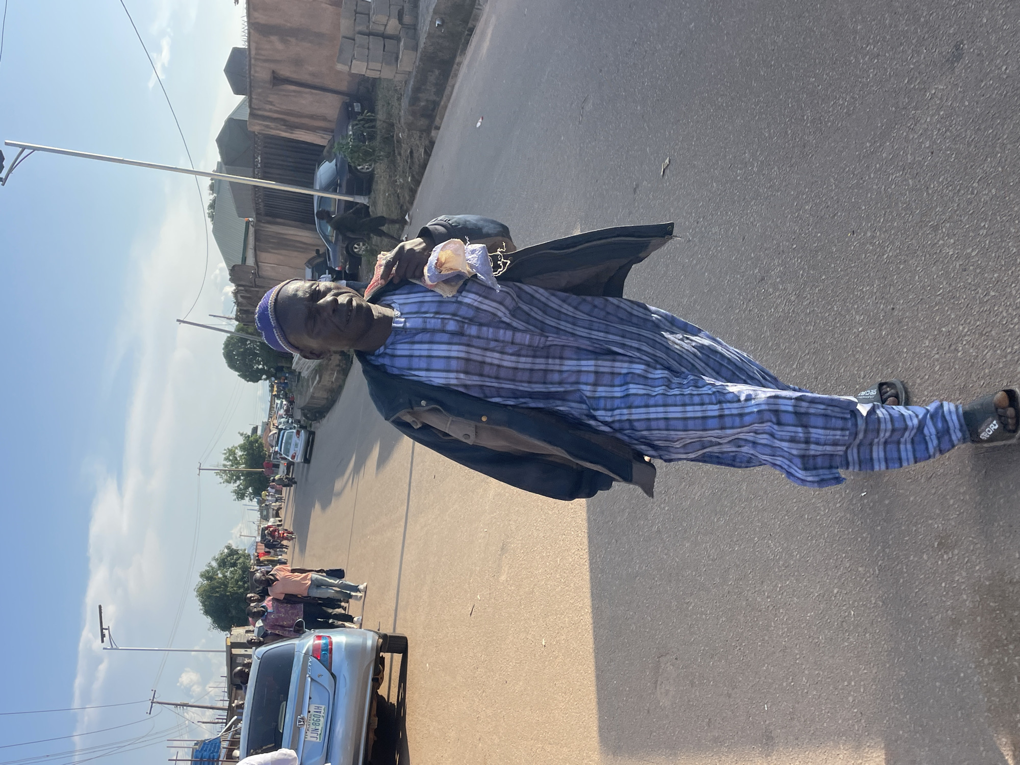 A man wearing a blue and white checkered tunic, a dark jacket, and a blue cap walks along a paved road in Gura Top, Nigeria. He is holding a wrapped item in his right hand. The road is lined with buildings, a wall, and trees. Other people and vehicles, including a blue car with license plate JJN-860AH, are visible in the background. The sky is blue with scattered clouds.