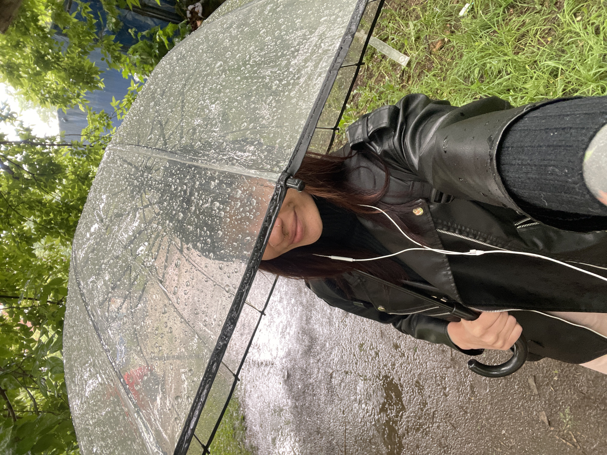 A person is holding a clear umbrella in a rainy outdoor environment. The person is wearing a black leather jacket and has brown hair. White wired headphones are visible. The ground appears wet, possibly a pavement or road. Greenery is visible in the background, including grass and trees. The location is likely Izhevsk, Russia, but this cannot be definitively confirmed from the image alone.