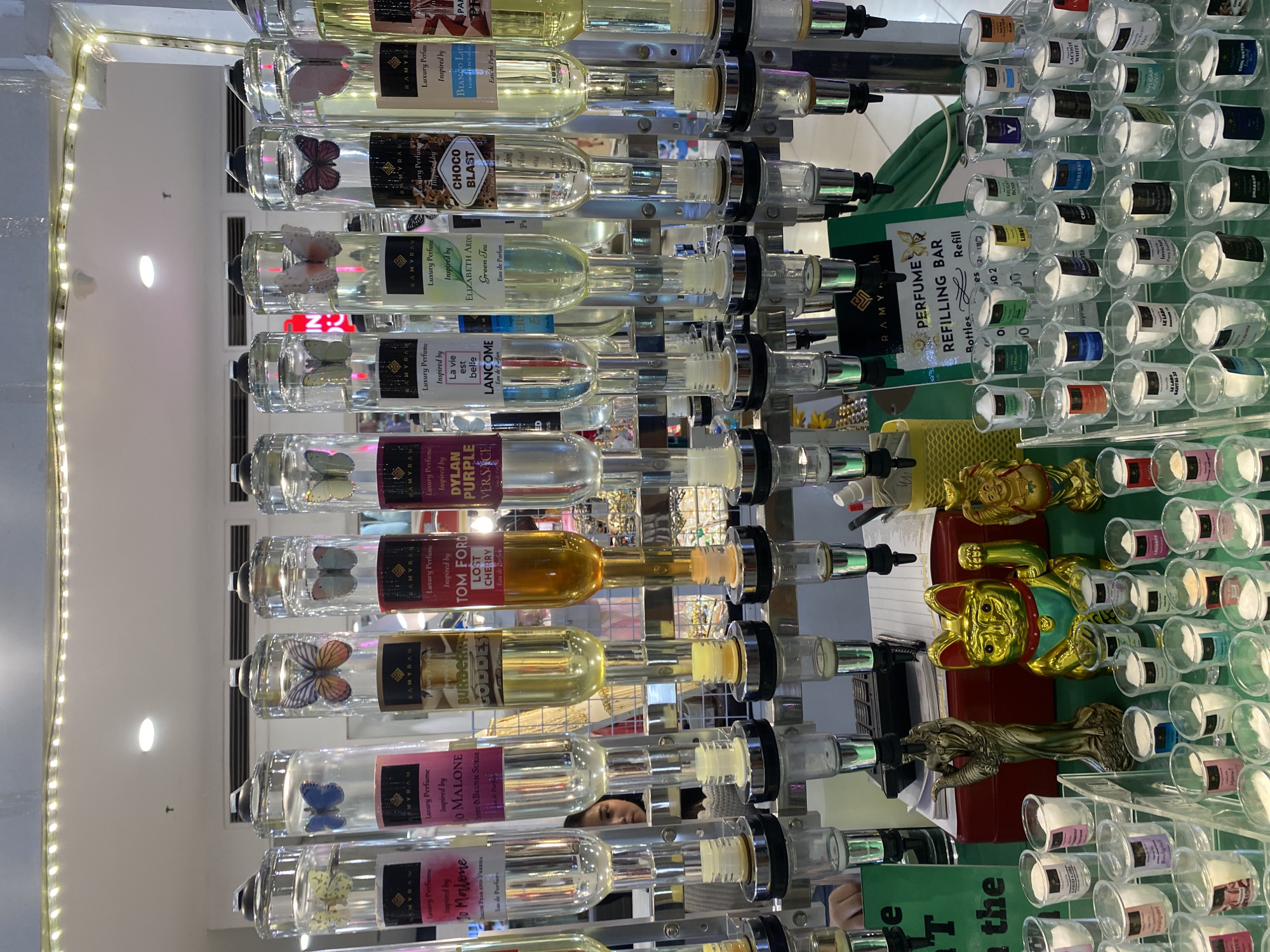 This image captures an indoor perfume refilling bar in what appears to be a retail setting, likely within a store or mall in Davao City, Philippines. The main subjects are numerous clear glass bottles, each containing a different colored liquid perfume, displayed in vertical rows on a multi-tiered stand. Many of these larger bottles feature decorative artificial butterflies inside.
Each large bottle is labeled "Luxury Perfume Inspired by" followed by a well-known fragrance brand or scent. Visible labels include: "BIANCO LA" (likely Bianco Latte), "CHOCO BLAST," "ELIZABETH ARDEN Green Tea," "LANCOME La vie est belle," "DYLAN PURPLE VERSACE," "TOM FORD LOST CHERRY," "BURBERRY GODDES" (Goddess), "Jo Malone Y&BLUSH SUEDE," and "Jo Malone PEAR AND FREESIA." The brand "RAMYRAM" is also visible on some labels.
To the right, a "PERFUME REFILLING BAR" sign is prominently displayed, indicating the service offered. Below this sign, numerous smaller, clear glass vials are arranged in horizontal rows, each with a white stopper and labeled with various fragrance names, likely representing different scents available for refill. Some identifiable names on these smaller vials include "LIBRE," "BOSS," "LE LABO SANTAL 33," "Invictur," "BURBERRY," "LACOSTE WHITE," "DRAKKAR," "BVLGARI AQVA," and "ONE."
The display includes decorative golden statues: a waving Maneki-neko (lucky cat) and a figure resembling a goddess or Lady Justice. An LED strip light illuminates the upper left edge of the display. The background is blurred but suggests a typical commercial interior. A subtle reflection of a person's face can be seen in one of the bottles. The time of day is not discernible, but the scene is brightly lit indoors.