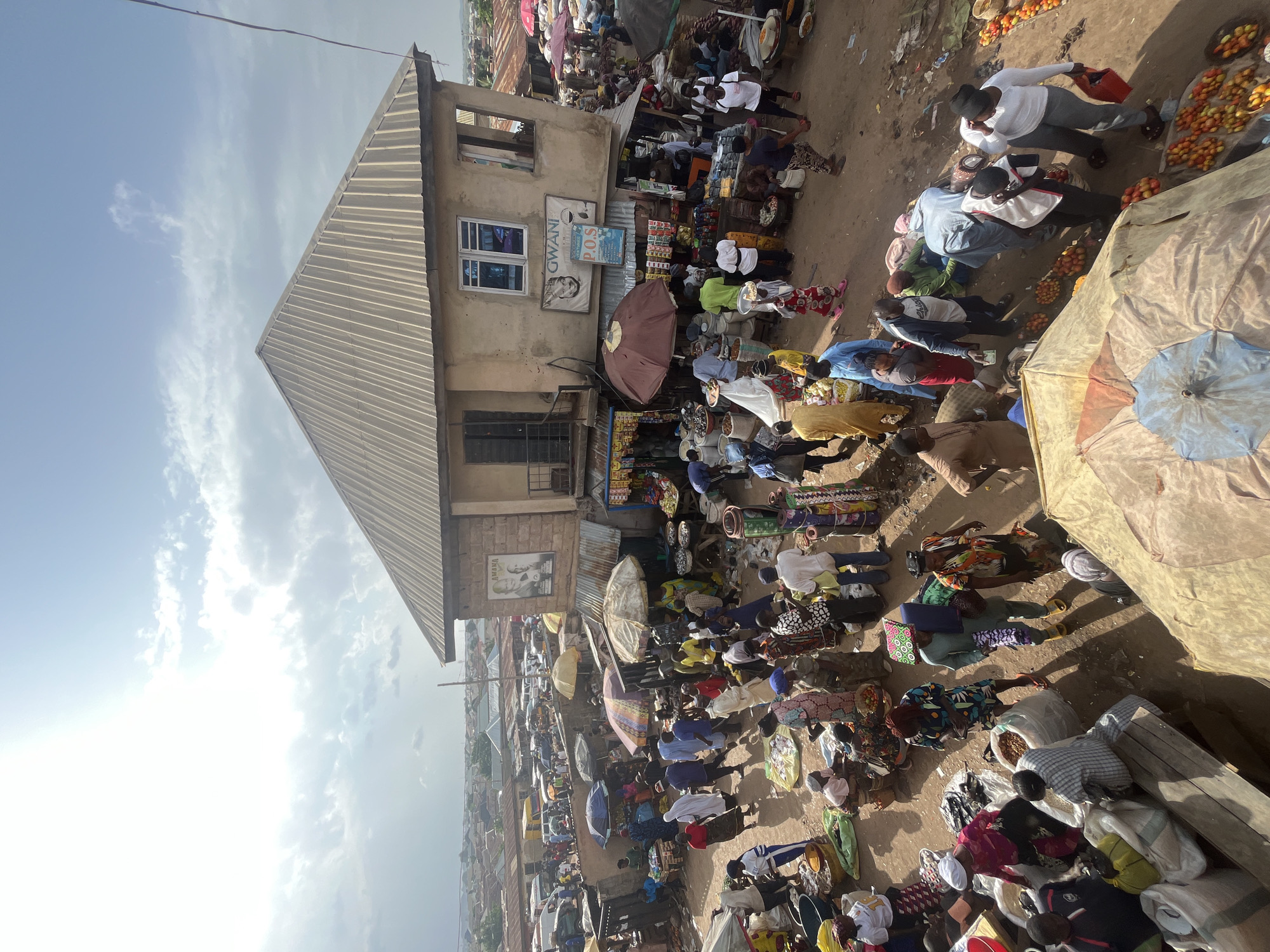 The image depicts a bustling outdoor market in Rafin Par, Nigeria. The scene is crowded with numerous people of various ages, dressed in diverse clothing, many of whom are engaged in commerce. Stalls are set up, displaying goods such as produce, grains, and textiles. Several vendors are interacting with customers, while others are attending to their merchandise. Umbrellas and awnings provide shade from the sun. In the background, more market activity and residential buildings are visible, extending into the distance. The overall impression is one of vibrant, active community life centered around trade.
