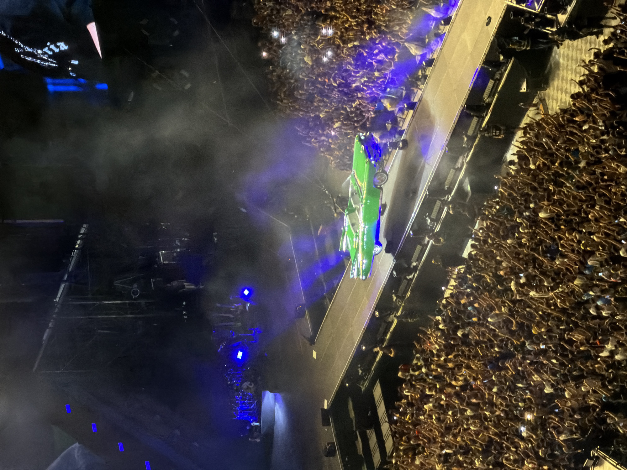The image depicts a concert with a large audience gathered. A bright green vintage convertible car is positioned on a raised platform or stage in the center, traversing a pathway. Smoke or haze is visible around the car and the stage area, illuminated by blue and white stage lights. The crowd, filling the lower portion of the frame, comprises many individuals with their arms raised, some holding up cell phones with their flashlights on, suggesting an active and engaged audience. The overall setting appears to be an indoor arena or a large venue designed for live performances. The specific location in Milan, Italy is not identifiable from the visual cues present in the image.