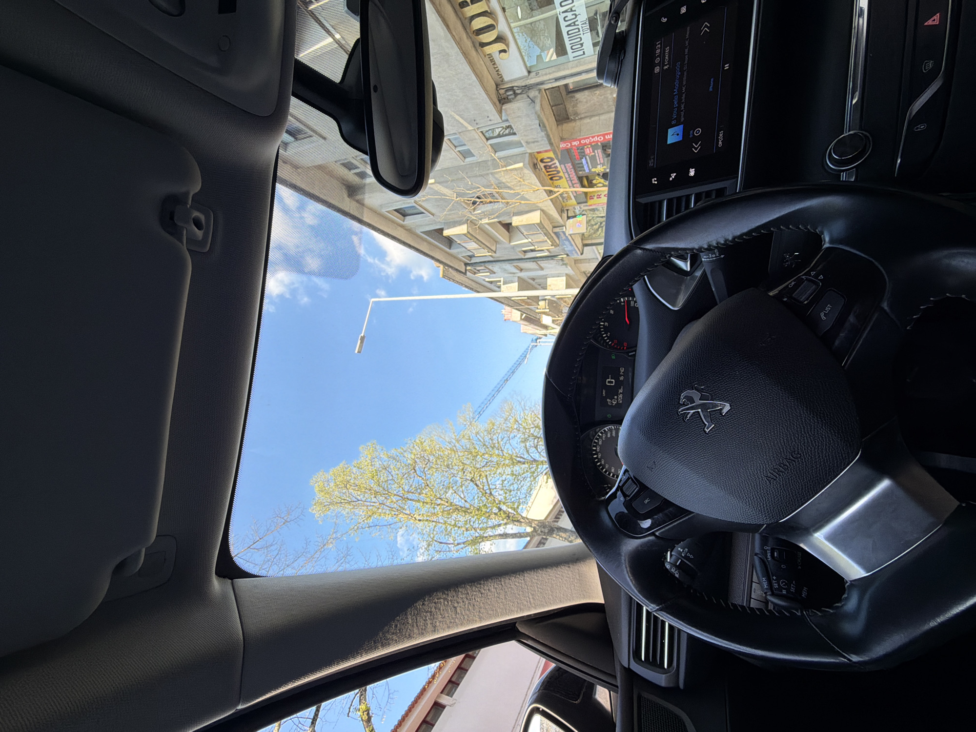 The image depicts a view from inside a car, likely a Peugeot, showing the steering wheel, dashboard display indicating "8 Vou pela Madrugada" by Mc Hilo, MC Wition, and others, and a portion of the interior. Through the windshield, the exterior scene shows buildings with signs including "SAPATARIA JOH", "LIQUIDAÇÃO TOTAL", and "Ouro", along with trees and a blue sky. OCR text indicates the location as "FONTES". Based on available information, the city is Vila Real, Portugal.