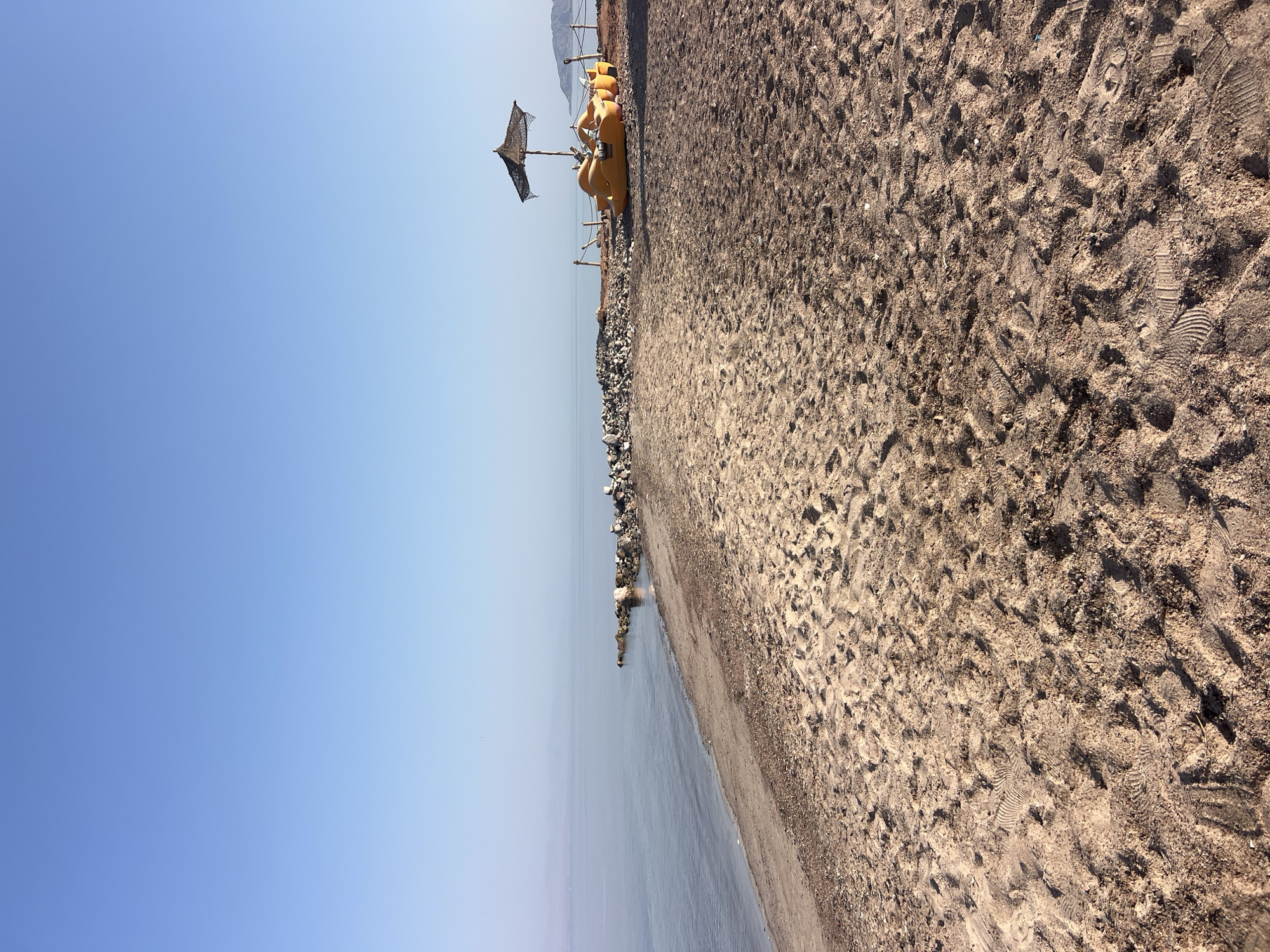 An outdoor, eye-level, full shot displays a sandy beach with calm, light blue water meeting the shore under a clear, pale blue sky. The sand is scattered with debris and footprints. To the right of the image, several yellow pedal boats are parked on the sand, some with a straw umbrella providing shade. The ocean stretches to the horizon, where distant, hazy mountains are visible. The scene is quiet and serene, suggesting a peaceful beach location. This beach is located in the city of Nuweiba, Egypt, a popular tourist destination known for its beautiful coastline and relaxed atmosphere.