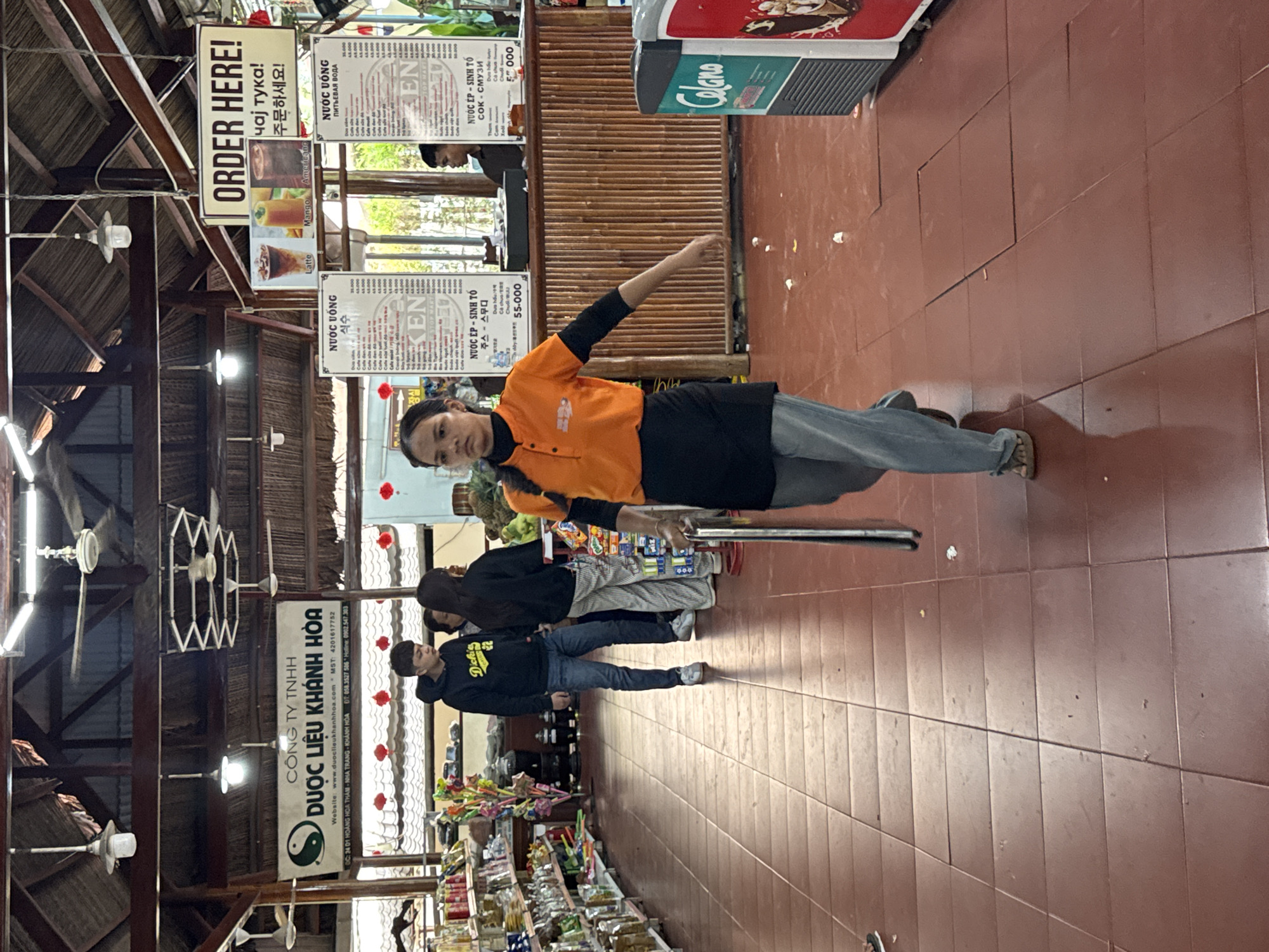 This image depicts the interior of a shop or restaurant in Nam Khanh Vinh, Vietnam, likely a food and beverage establishment given the menus and beverage cooler. A young woman, wearing an orange shirt and black apron, walks forward across a tiled floor. She is holding a metal pole in her right hand and appears to be in the process of cleaning or tidying the area, as evidenced by small pieces of debris on the floor. In the background, several other individuals are visible, some interacting near a counter. Shelves stocked with various products line the left side of the frame. A prominent sign above the counter reads "ORDER HERE!" in English, with additional text in what appears to be Russian and Korean, suggesting an international clientele. Menu boards displaying drink options are also present. The overall environment suggests a casual, possibly tourist-oriented establishment with a rustic aesthetic, indicated by the wooden counter and ceiling structure.