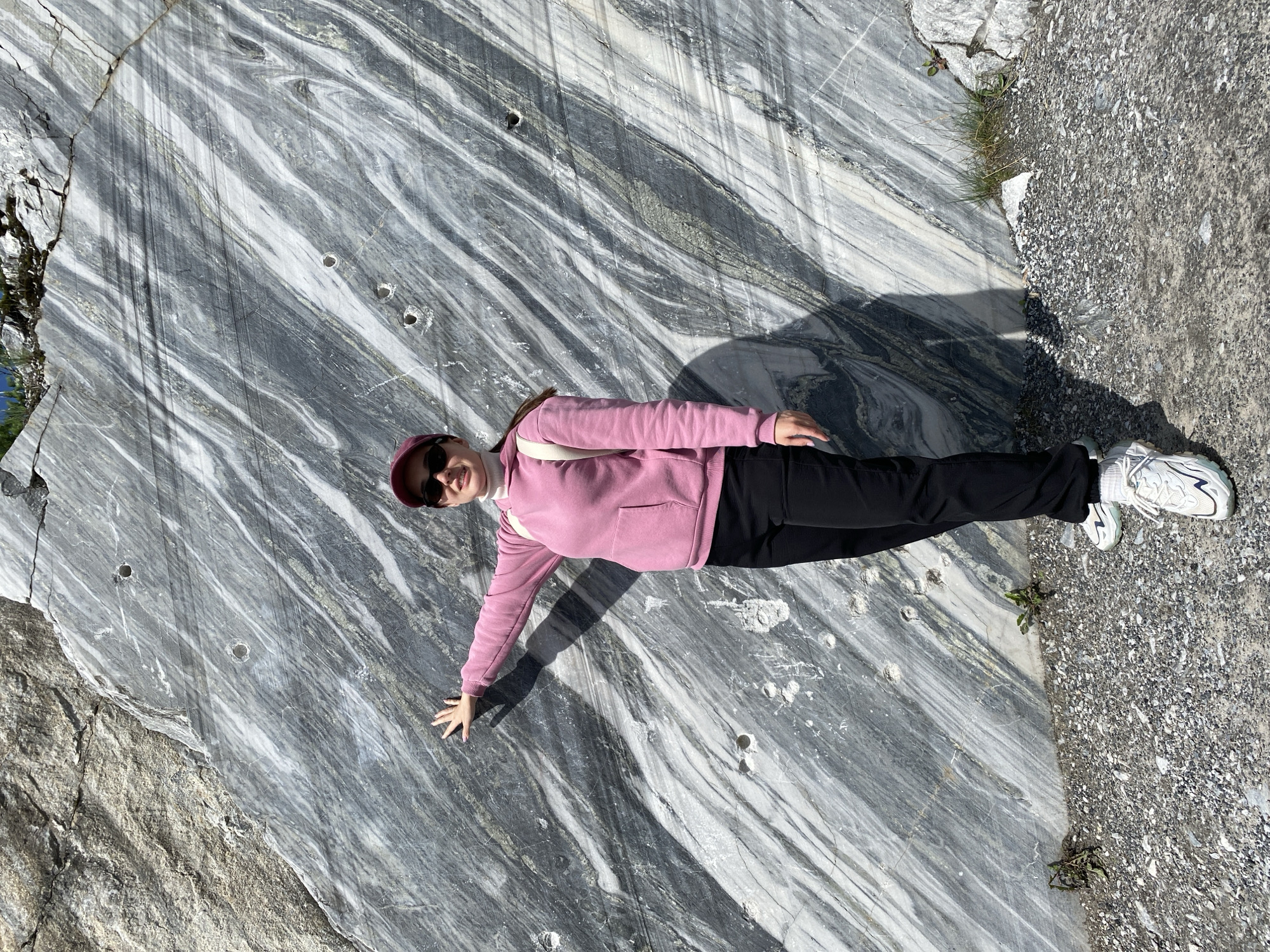 In Ruskeala, Russia, a woman stands in front of a large, striated marble cliff face. The marble exhibits a dynamic pattern of gray, white, and black veins, indicative of its geological origin. The woman, wearing a pink hoodie, black pants, a maroon baseball cap, and sunglasses, is positioned to the left of the frame, with her left arm extended and hand touching the rock face. She has a white backpack slung over her right shoulder. Her shadow is cast diagonally across the marble. The ground in front of the cliff is covered with gravel and small rocks, with some sparse vegetation growing near the base of the rock. Several circular indentations are visible on the marble surface, possibly from past quarrying activities. The scene is illuminated by bright sunlight, creating strong contrasts and shadows.