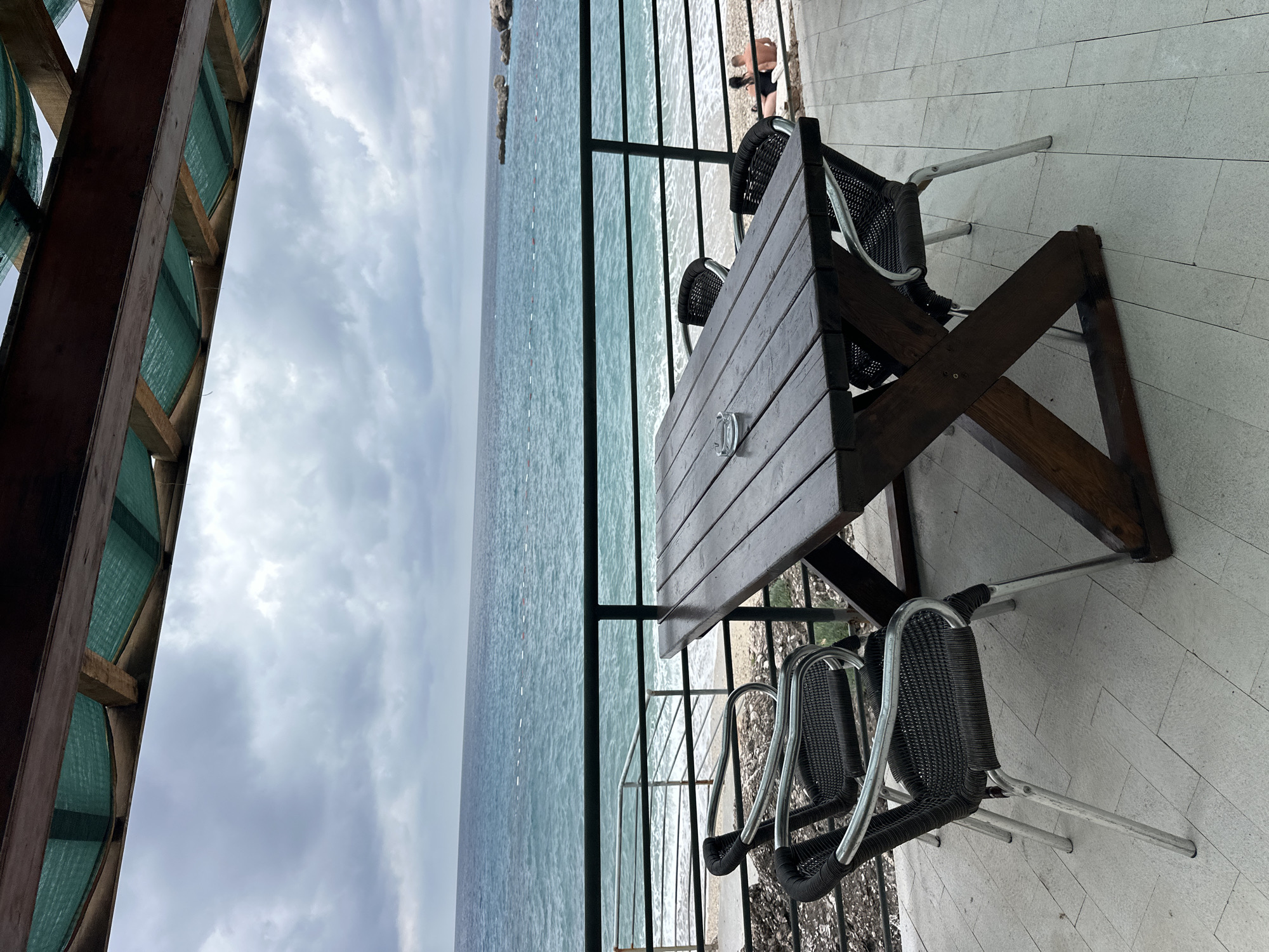This media file captures an outdoor dining or seating area on a terrace overlooking the sea, likely in Drobnići, Montenegro. In the foreground, a sturdy dark wooden table with a slatted top is set with a clear glass ashtray. Four chairs with black woven seats and backs, supported by silver-colored metal frames, are arranged around the table. The terrace floor is made of light-colored, rectangular tiles.
A dark metal railing separates the terrace from the area below, which consists of a pebbly beach. On the beach, two individuals are partially visible. One person is bending over, wearing dark shorts, and another's legs are seen further along the shore. Beyond the beach, the sea stretches out to the horizon, displaying shades of blue and green. White dashed lines, possibly indicating buoys or a swimming area, are visible in the water. A rocky coastline extends into the sea on the far left.
Above the scene on the left, part of an overhead structure, possibly a pergola or a building's overhang, is visible. It features dark wooden beams with light green, semi-transparent mesh or fabric panels. The sky above is overcast, filled with grey clouds, suggesting a cloudy day. There is no visible text in the image. The overall atmosphere is calm and tranquil, typical of a coastal setting.