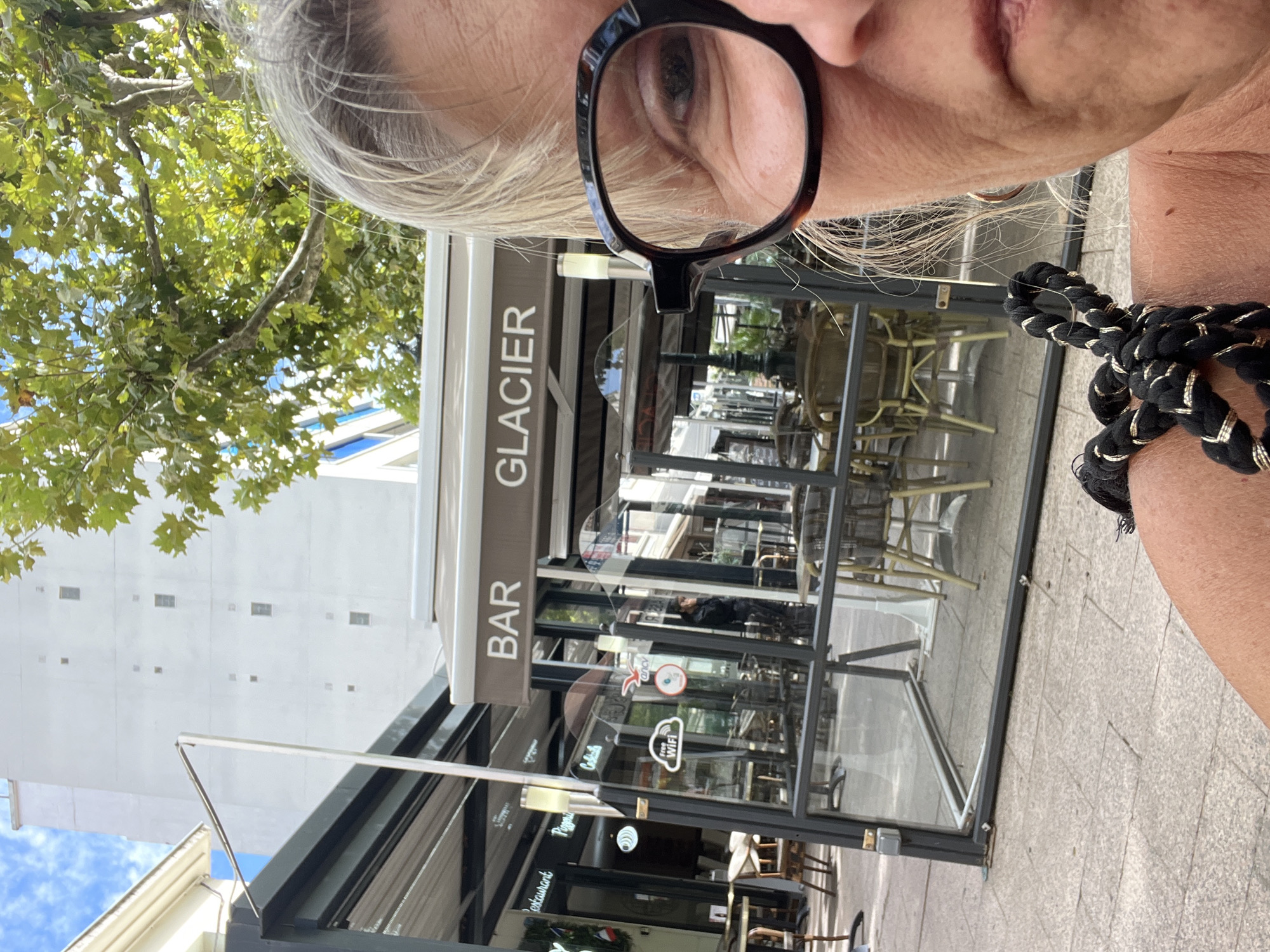 The image shows a close-up of a woman's face and shoulder, wearing glasses. In the background, a building with windows and a tree with green leaves are visible. There is also a sign that reads "BAR GLACIER". The scene appears to be an outdoor seating area of a cafe or restaurant. The woman is positioned in the foreground, with her face and part of her shoulder taking up the majority of the frame. The background is blurred, suggesting a shallow depth of field. The lighting is bright, indicating it is daytime. The location is identified as Montélimar, France, which is a city in the Drôme department in southeastern France.