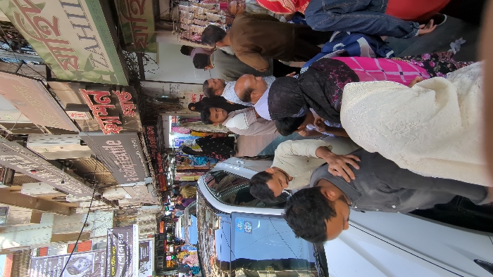 The image displays a densely crowded commercial street in Dhaka, Bangladesh, under clear daylight conditions. Multi-story buildings with numerous retail establishments flank both sides of the narrow thoroughfare. Shop signs featuring English text such as "Rafiq Textile" and "ZAHID," along with extensive Bengali script, are prominent. Banners, some displaying images of individuals and Bengali text, including one mentioning "শোকাহত" (grieving/mourning), are suspended overhead across the street.
The street is heavily populated by numerous individuals, predominantly adults. People are observed walking, standing, and congregating, with significant density of movement. Attire includes traditional South Asian garments, with women wearing headscarves and men in kurta-style shirts, alongside some Western casual wear.
A white SUV is positioned in the center-left foreground, partially visible, with its front oriented towards the background. Individuals are closely navigating around the vehicle. A man in a grey shirt is seen leaning forward near the SUV, while another man in a light-colored shirt stands adjacent to him. In the immediate foreground, the back of a woman's head covered by a white, textured headscarf is visible. The overall scene depicts a bustling urban market environment.