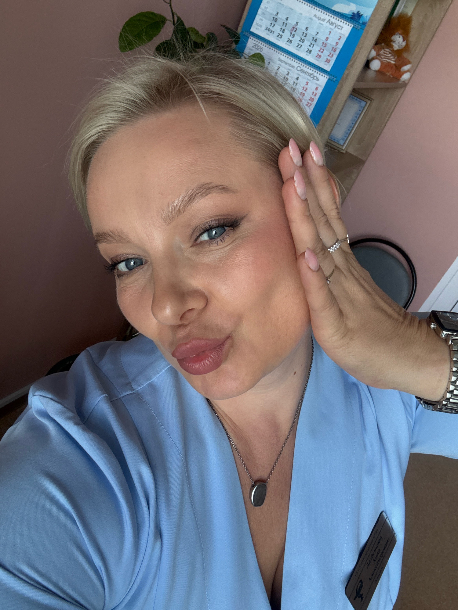 A woman with blonde hair and blue eyes is taking a selfie in what appears to be an office or medical setting. She is wearing a light blue scrub top, a silver pendant necklace, and a watch on her left wrist. Her right hand is touching her face. Behind her, a calendar showing August and September is visible, along with a small stuffed lion toy on a shelf. The wall behind her is pink. The overall setting is indoors. There is no indication of the city of Slawharad, Belarus in the image.
