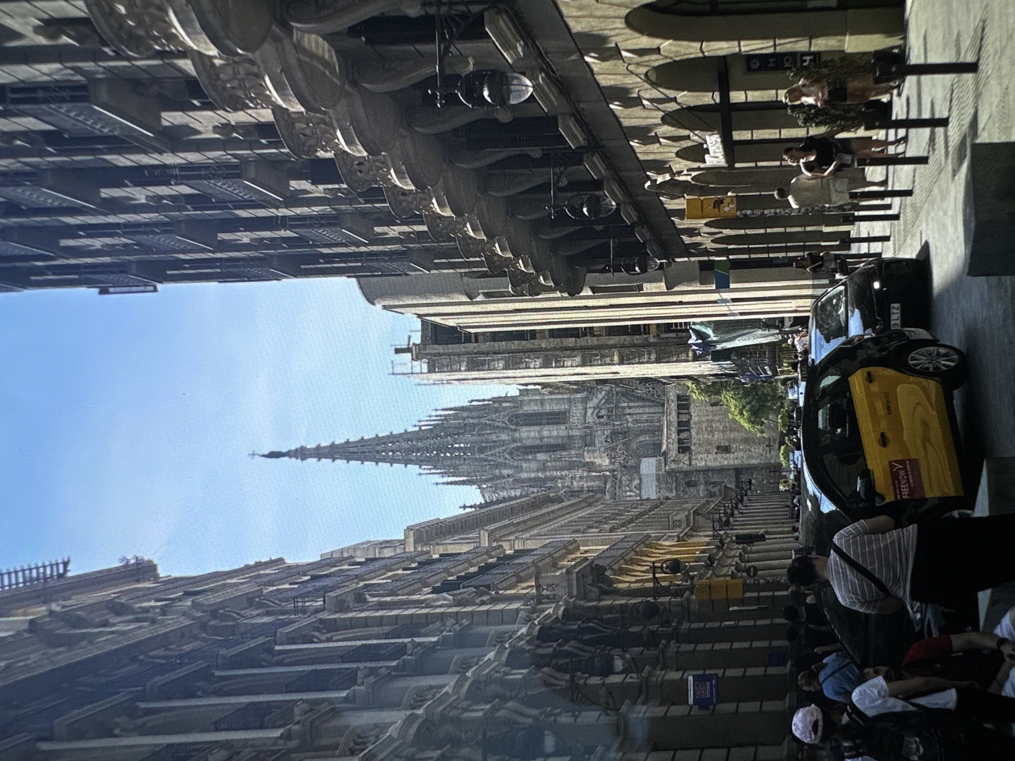 The image shows a street scene in Barcelona, Spain, with the Cathedral of Barcelona visible in the distance. The street is lined with tall buildings with ornate facades. People are walking on the sidewalk, and there are parked cars, including a taxi. The sky is clear and blue.
