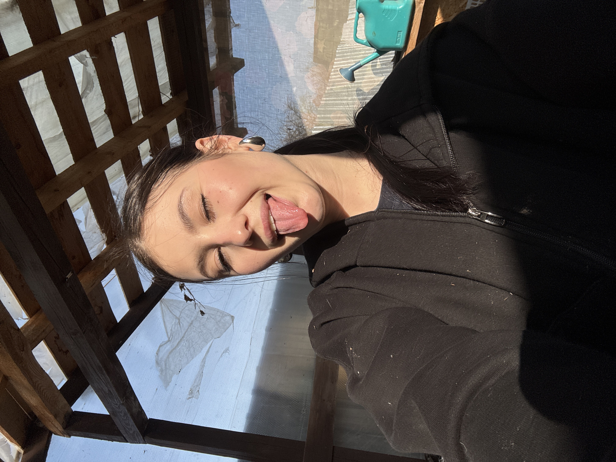 A woman with dark hair pulled back is sticking out her tongue, with her eyes closed, wearing a dark hooded jacket in a structure made of light wood and plastic sheeting. The location is likely not Фоки, Russia, since the image provides no indication of location and this information cannot be determined from the available visual data. A watering can is visible through the plastic sheeting. The structure appears to be a small greenhouse or shed.