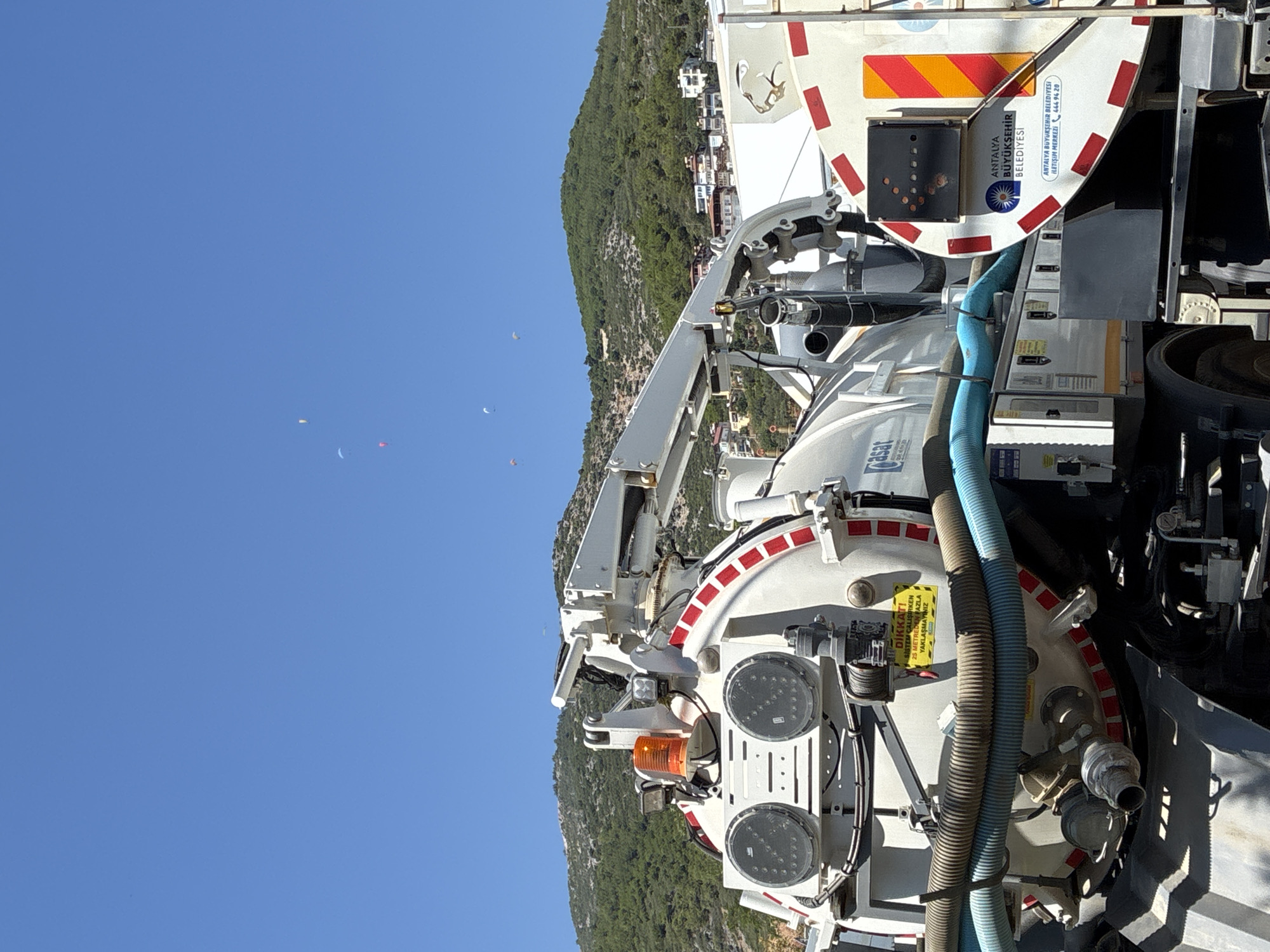 In Alanya, Turkey, a large white sanitation truck is parked on a sunny day. The truck is equipped with a large tank, hoses, and a boom arm, suggesting it is used for waste collection or maintenance. In the background, a lush green mountain slopes upwards, dotted with buildings and houses. The sky above is a clear, vibrant blue. Several paragliders can be seen in the distance, suspended in the air against the sky, adding a recreational element to the scene. The truck features several labels, including one in Turkish that reads "DİKKAT! SİSTEM ÇALIŞIRKEN 25 METREDEN FAZLA YAKLAŞMAYINIZ" which translates to "CAUTION! DO NOT APPROACH WITHIN 25 METERS WHILE THE SYSTEM IS OPERATING." Another label indicates "ANTALYA BÜYÜKŞEHİR BELEDİYESİ," identifying the truck as belonging to the Antalya Metropolitan Municipality.