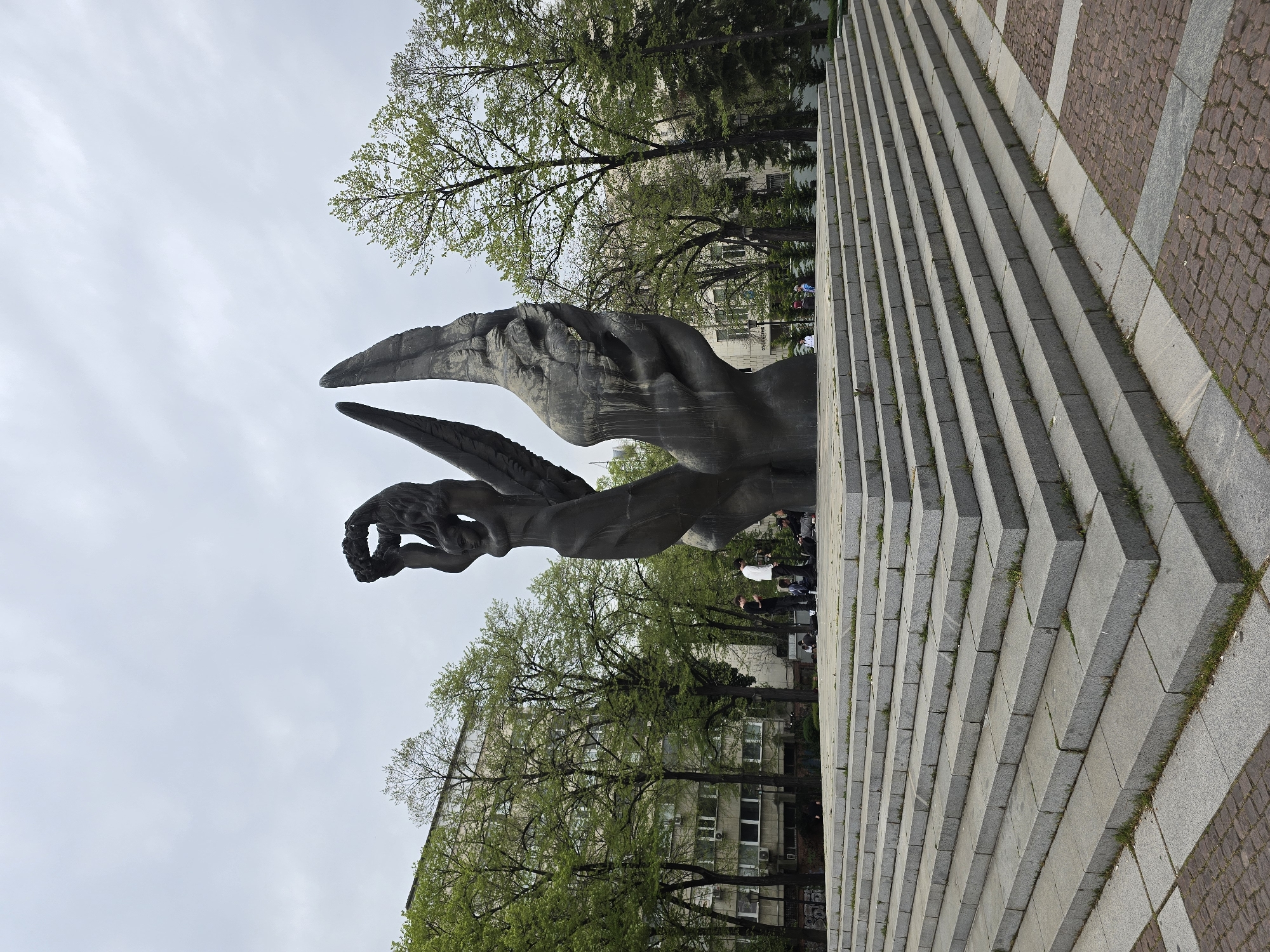 The image displays a large, dark monument featuring a winged, human-like figure holding a wreath, set against an overcast sky. The monument, appearing to be made of dark bronze or stone, is positioned in the left-center of the frame, with its large wings sweeping upwards.
To the right, a long, wide flight of light-colored stone steps ascends diagonally from the foreground. The ground adjacent to these steps is paved with a mix of rectangular stone slabs and sections of cobblestones, with small patches of grass growing between the cracks.
In the mid-ground and background, several trees with fresh green leaves, some still sparse, frame the scene. Behind the trees, a multi-story building with numerous windows and visible air conditioning units suggests an urban setting. A few people are scattered in the mid-ground near the base of the monument and the building, appearing to be casually present in a public space.
The time of day is daytime, under an overcast sky that provides soft, diffused lighting. There are no harsh shadows. Faintly visible on the side of the building in the background is the partial Cyrillic word "Обидчивы." The overall scene suggests a public park, square, or memorial site in a city.