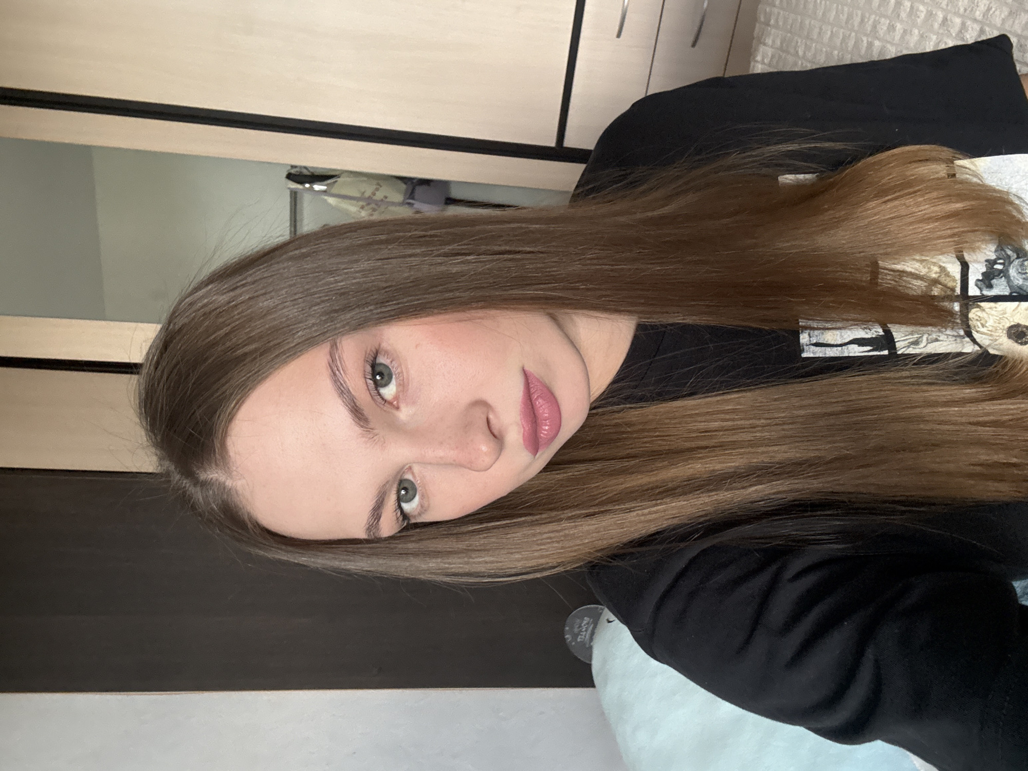 The image shows a young woman with long, brown hair, fair skin, and green eyes. She is wearing a black t-shirt. She is looking directly at the camera, with her head tilted slightly to the right. Her lips are painted with a pink lipstick. In the background, there is a dark wooden wardrobe and a light-colored wall. A pale blue blanket is visible on the left side of the image, and a white textured surface, possibly a bedspread, is on the right. The overall lighting suggests an indoor setting, likely a bedroom. The image does not contain any explicit indicators of the specific location within the city of Perm, Russia.