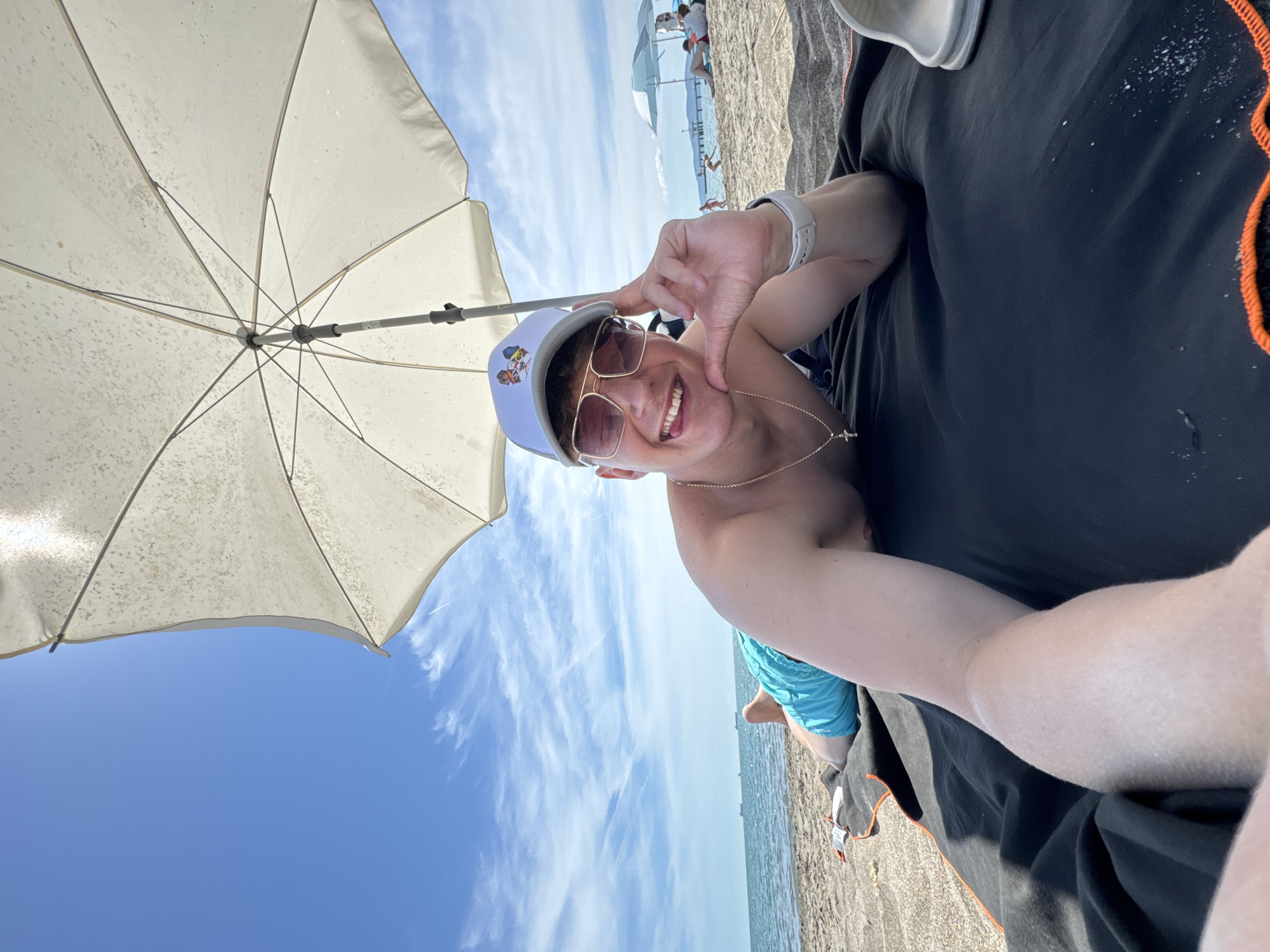 A man is lying on a beach towel on the sand, facing the camera. He is wearing blue swim trunks, a white baseball cap with a graphic on the front, and sunglasses. A gold chain with a cross pendant is visible around his neck. He is smiling and sticking his tongue out slightly, with his right hand resting on his chin. He is positioned under a large, white beach umbrella. In the background, the sea is visible, with several boats in the distance. Other beachgoers and umbrellas are also visible further down the beach. The sky is blue with scattered white clouds. The location appears to be a beach in Burgas, Bulgaria.