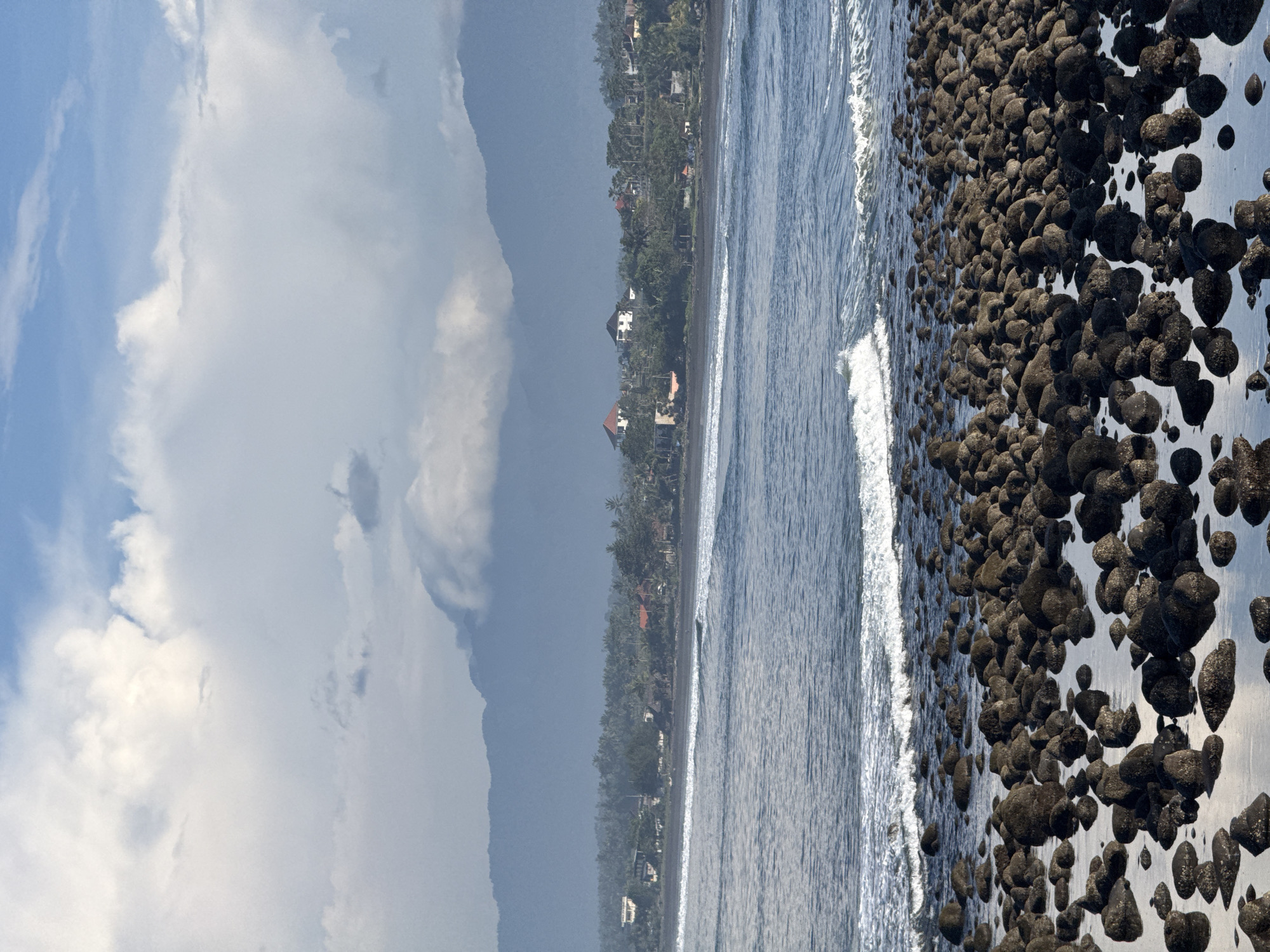 The image depicts a beach scene in an unidentified location. The foreground features a rocky shoreline with numerous dark, rounded stones. The water's edge is visible as a line of white foam from the crashing waves, separating the stones from the ocean. Beyond the waves, the ocean extends towards the horizon. A coastline with dense vegetation and scattered buildings stretches along the edge of the ocean. The sky is partially cloudy with both bright blue patches and darker, overcast areas.