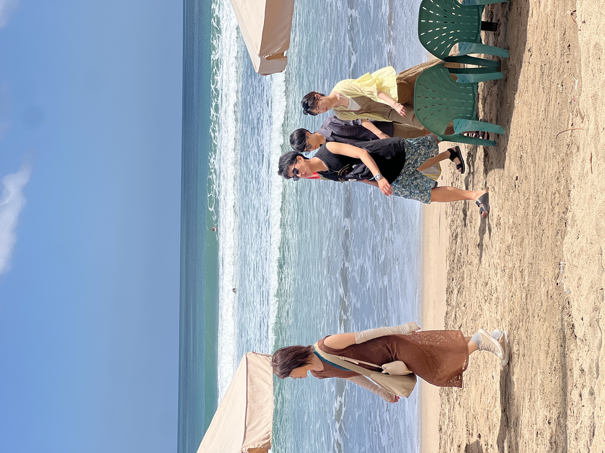 The image depicts a sunny day on a sandy beach, likely in Kuta, Indonesia, given the presence of a surfboard in the background. Four individuals are walking on the shore. On the left, a woman wearing a brown dress and arm sleeves walks away from the camera, carrying a tan shoulder bag. To her right, a group of three people is approaching. The first person in this group, a male, wears a black sleeveless shirt and patterned shorts. Next to him is another male in a grey button-down shirt, followed by a female in a yellow cardigan over a white top and brown pants. In the foreground, two green plastic chairs are positioned on the sand. The ocean is visible in the background, with waves breaking on the shore. Two beach umbrellas, one on the left and another on the right, provide shade.