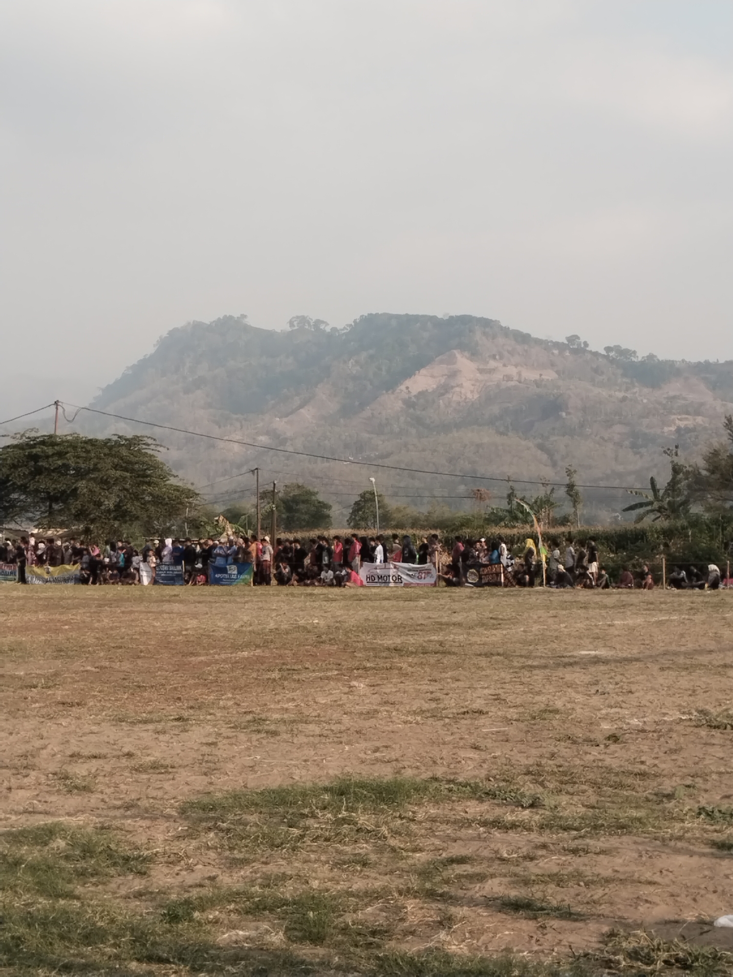 In Margasari, Indonesia, a crowd of people is gathered on a dry, grassy field in front of a backdrop of hills. The people are standing and sitting, some holding banners. The banners appear to be promotional, with text such as "HD MOTOR". The environment is hazy, suggesting a dusty or smoggy atmosphere. The overall scene depicts an outdoor event or gathering.