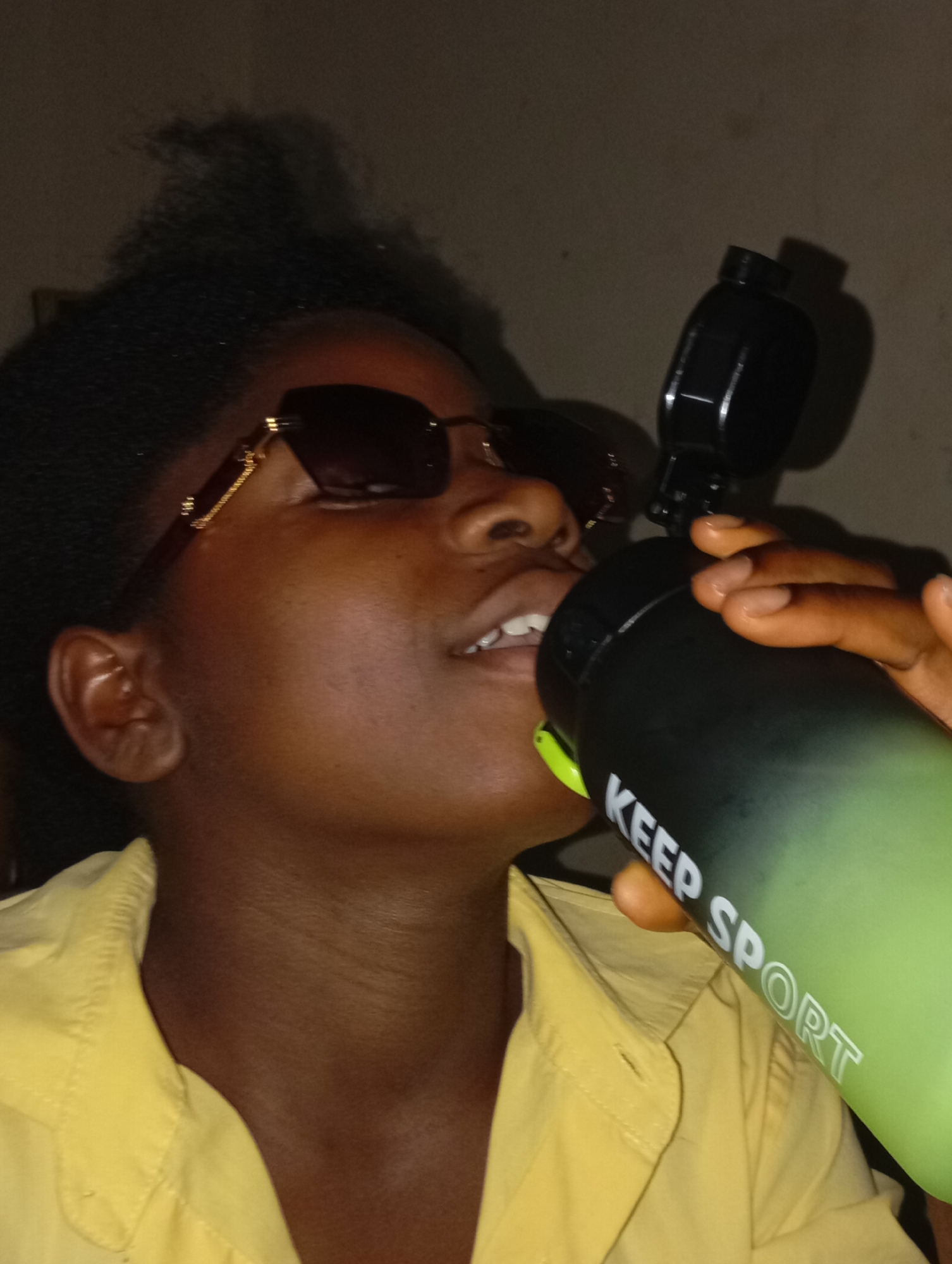 A person with dark skin and short, curly dark hair is drinking from a water bottle with "KEEP SPORT" printed on it. They are wearing brown sunglasses with gold detailing and a yellow button-up shirt. The background appears to be a dimly lit wall. The location of the photo in Jos, Nigeria, cannot be determined.