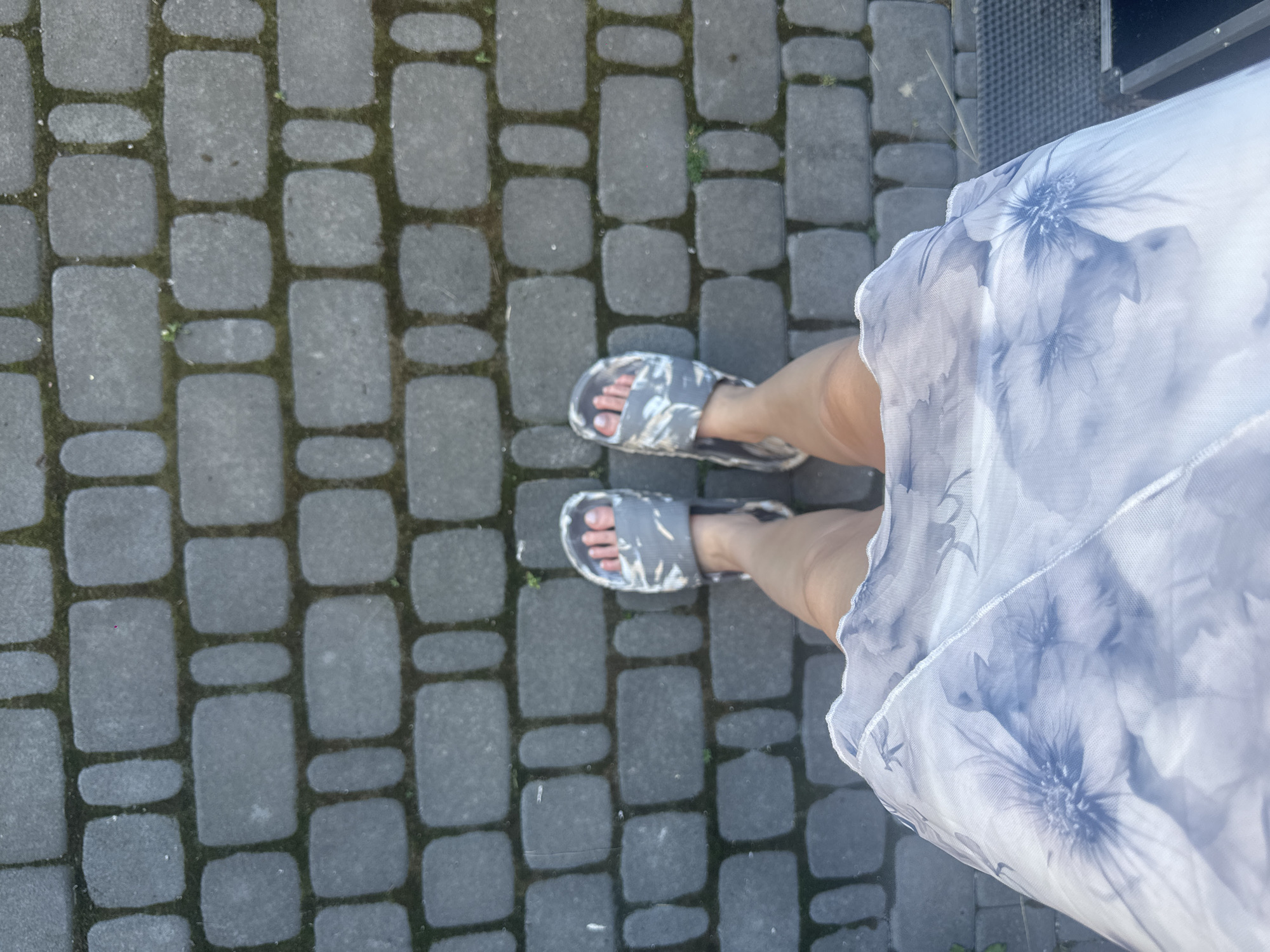 A first-person perspective shot shows a person wearing gray patterned slides and a light blue floral sheer dress standing on a gray brick pathway. The pathway is laid in a pattern of rectangular and square bricks with visible green moss growing between them. The person's legs are visible from the knees down, with the hem of the dress falling around the thighs. In the bottom right corner, a dark gray textured surface, possibly a doormat, is visible. The overall scene is outdoors on a paved surface.