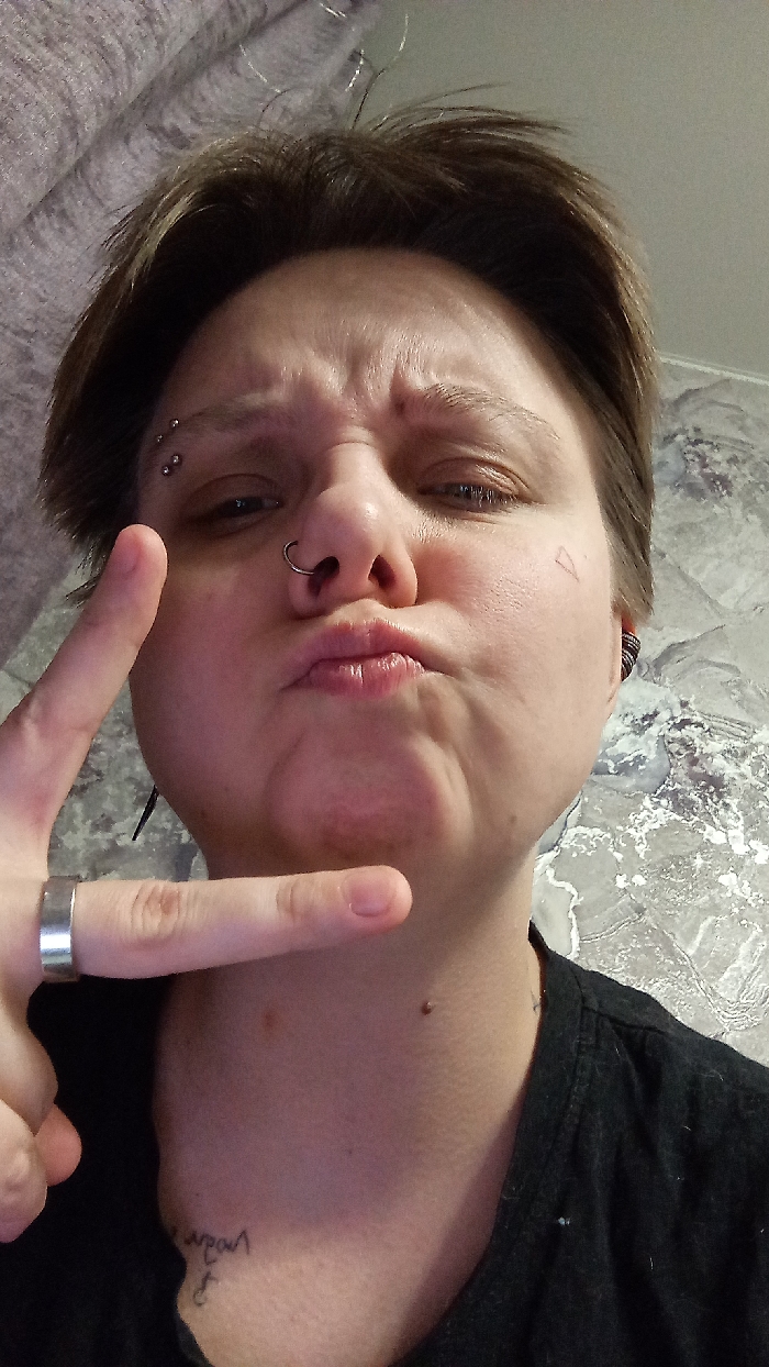 The image displays a close-up, head-and-shoulders portrait of an individual, gender indeterminate, with short, light brown hair, captured indoors in Saratov, Russia. The subject is directly facing the camera with a pouting expression, characterized by pursed lips, a scrunched nose, and furrowed eyebrows.
The individual has three small, metallic stud piercings vertically aligned above the left eyebrow and a metallic septum ring in the nose. A faint, triangular outline tattoo is visible on the right cheek. A dark earbud or similar device is present in the right ear. The left hand is raised, performing a two-finger "V" gesture, and a plain metallic band ring is worn on the ring finger. On the left side of the neck, a handwritten script tattoo, partially legible as 'любовь' (love), is visible near the collarbone, along with a small, dark mole. The subject is wearing a dark-colored garment, possibly a t-shirt, visible at the neckline and shoulder.
The background consists of a light-colored wallpaper featuring a subtle, abstract grey pattern. The upper left corner shows a blurred grey textile, possibly a curtain or bedding.