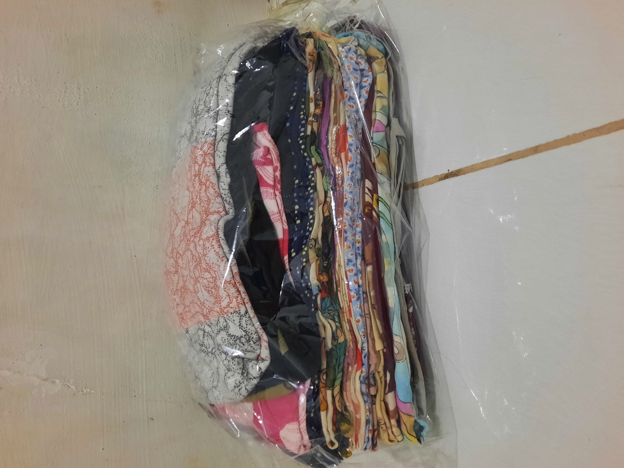 The image shows a stack of folded clothes wrapped in clear plastic. The clothes are a variety of colors and patterns, including floral prints, abstract designs, and solid colors. They are neatly folded and piled on top of each other, creating a somewhat uneven stack. The plastic wrap is slightly crinkled, indicating that the bundle has been handled. The background is a plain, textured wall that appears to be painted. The floor beneath the bundle is tiled, with a visible grout line running diagonally across the image. The lighting is even, with no strong shadows, suggesting an indoor setting. Based on the provided information, the location is Lhokseumawe, Indonesia.