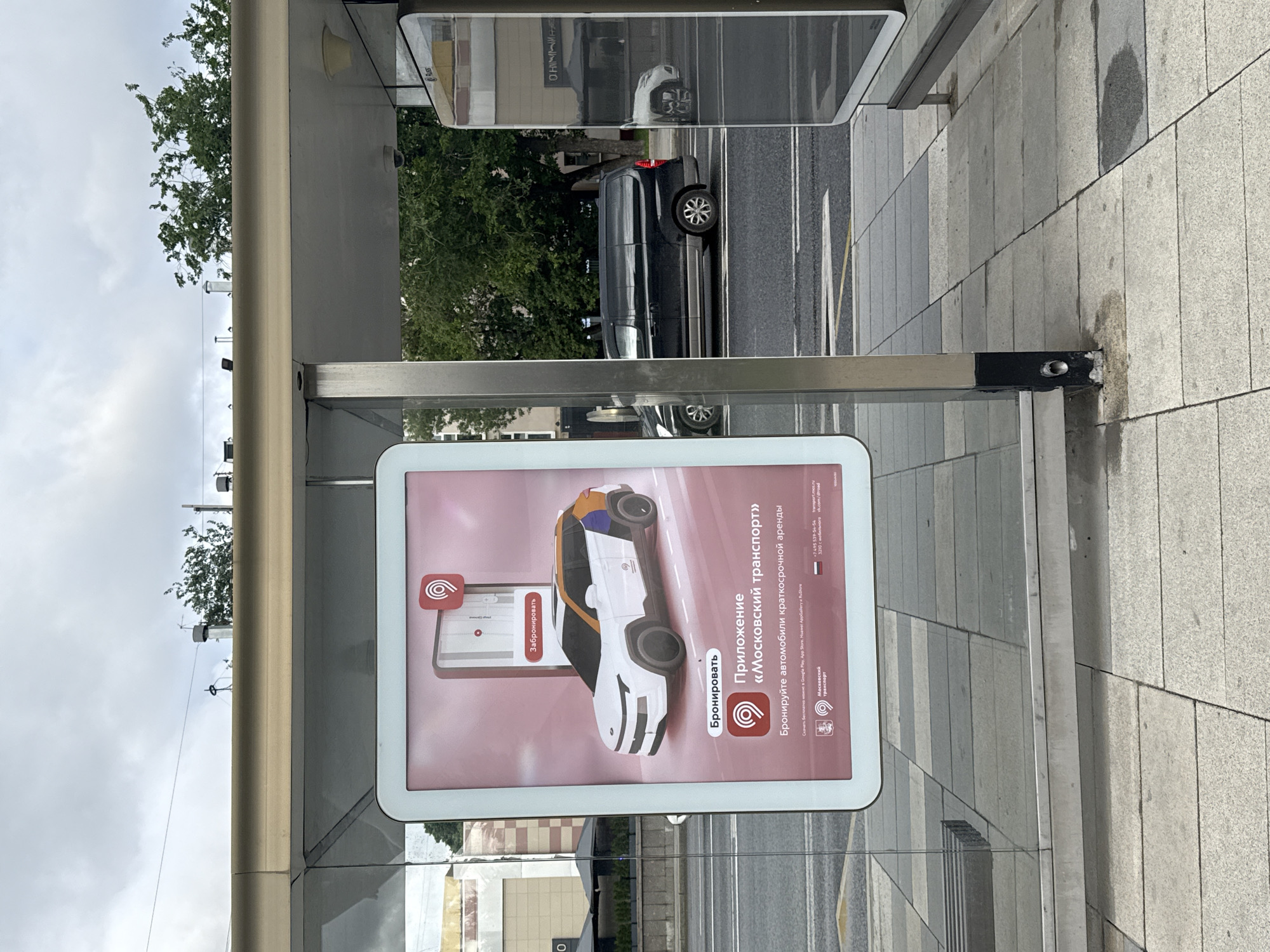 This image shows a bus stop in Moscow, Russia. The bus stop has a sign advertising the "Moscow Transport" app for renting cars. A street and sidewalks are visible behind the bus stop. Cars are driving on the street.