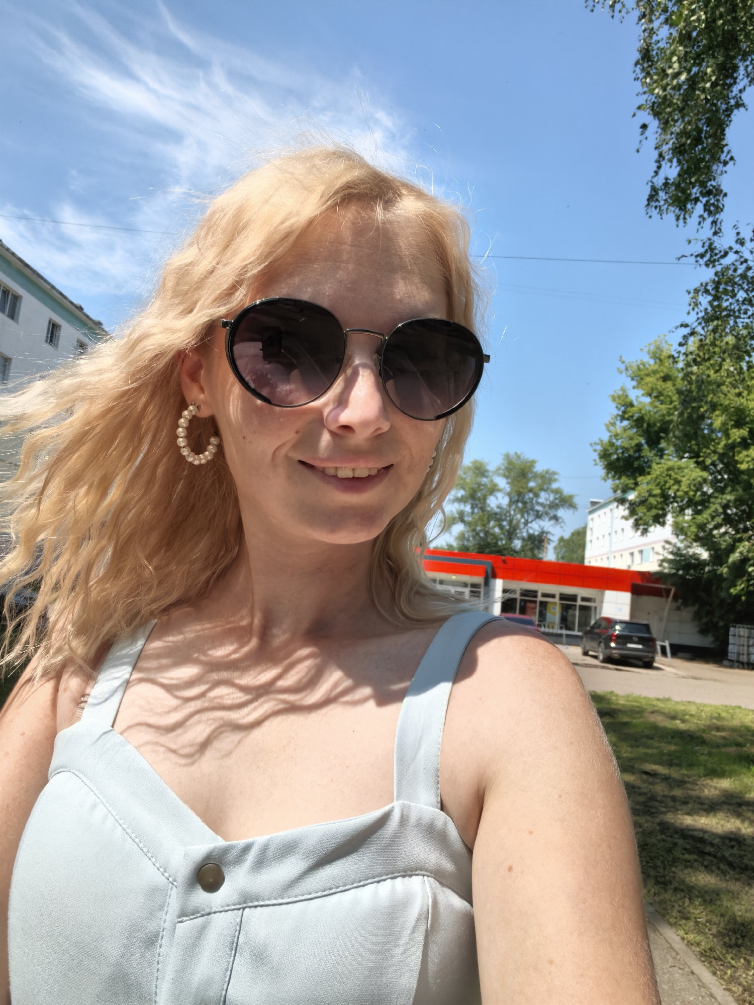 A blonde woman wearing sunglasses and pearl hoop earrings takes a selfie outdoors. She is wearing a light blue dress. Behind her, a modern building with red accents can be seen, along with trees and a parked car. The sky is bright blue with some white clouds. The image was taken on the territory of "Magnit" supermarket in the city of Sterlitamak, Russia.