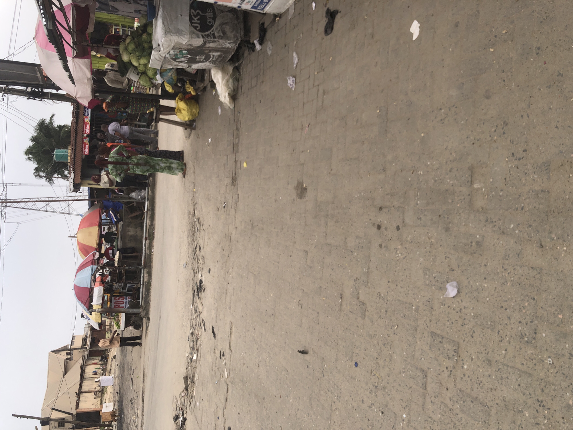The image depicts a street scene in Ado, Nigeria, characterized by a busy outdoor market. Several stalls are set up, covered by colorful umbrellas, displaying various goods. In the foreground, the ground is paved with concrete blocks, littered with debris, including plastic bags, paper, and other discarded items.
On the left side of the image, a man in a light brown shirt and dark pants stands near a stall with a sign that reads "POS." He appears to be interacting with the stall. Further left, a building with a light-colored, sloped roof is visible, along with a dog.
In the center and right portions of the image, more market stalls are present. People are seen standing and walking around the stalls. One stall prominently features a large pile of green coconuts. A person in a patterned green dress is standing near this stall. Several plastic bags and other items are scattered on the ground near the stalls. A large, grey container or vehicle with signage that includes "JK3 grtis" is parked on the right side. Overhead, power lines and utility poles are visible, indicating an urban environment. The sky is overcast.