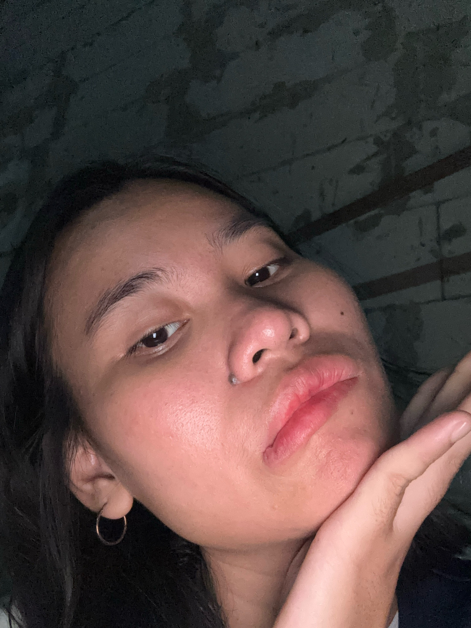 The image shows a close-up, low-angle selfie of a young woman. She is resting her right hand on her chin, with her fingers splayed. Her lips are slightly pursed, and she has a small stud piercing on her philtrum. She is wearing a hoop earring in her left ear. Her skin appears to have a subtle sheen. The background is dark and appears to be a textured wall, possibly concrete or brick, with some horizontal lines suggesting construction or a weathered surface. The lighting is dim, casting shadows on her face and highlighting certain features. There is no indication of her specific location within East Jakarta, Indonesia, beyond the inferred setting of an indoor or sheltered area with a rough wall.